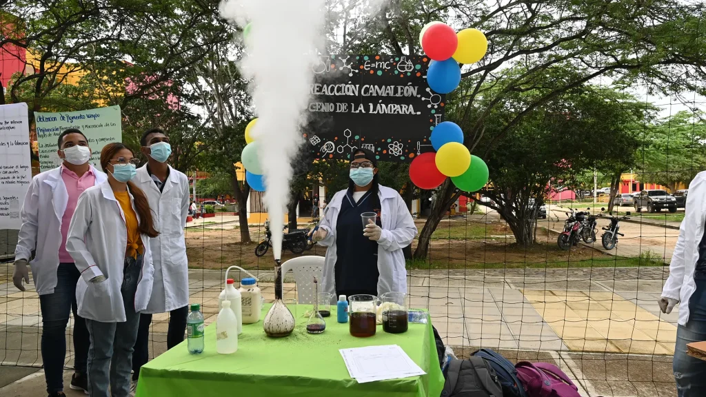 Grupo de estudiantes con batas de laboratorio y mascarillas realiza un experimento químico al aire libre. Sobre una mesa cubierta con un mantel verde hay frascos, envases y una botella de laboratorio de la que emerge una columna alta de humo blanco. Detrás, un cartel decorado con dibujos científicos y globos de colores anuncia la actividad. Se observan árboles, motocicletas y edificios en el fondo del campus.
