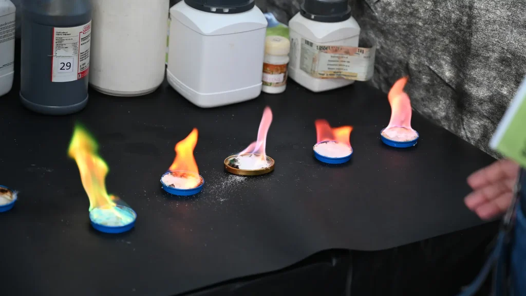 Varias tapas metálicas alineadas sobre una mesa negra sostienen pequeñas cantidades de sustancias químicas que arden con llamas de diferentes colores —verde, naranja, rosa y rojo— como parte de una demostración química. Al fondo se observan frascos y recipientes de laboratorio.