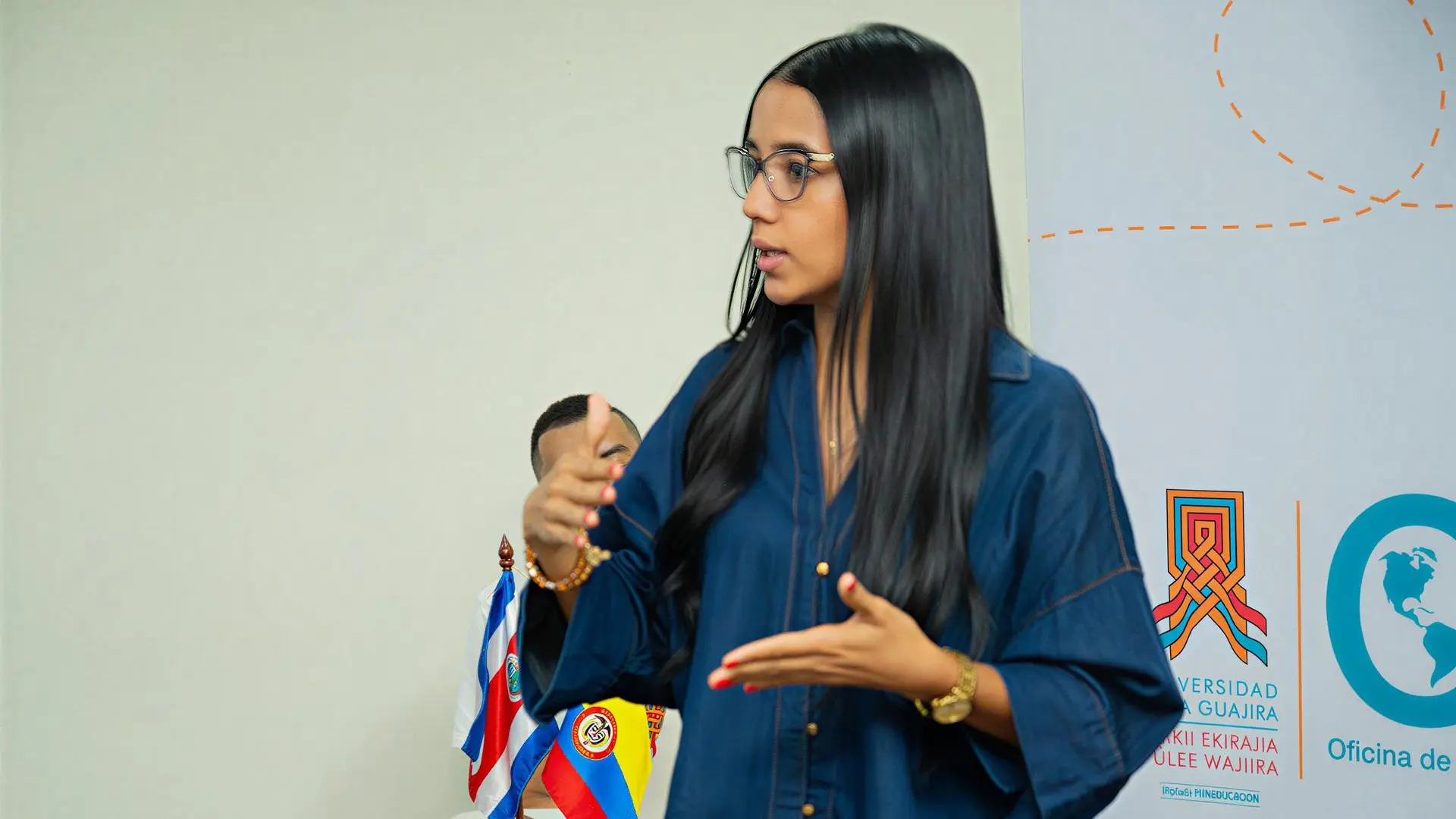 Joven profesional realizando una exposición en un evento institucional relacionado con internacionalización y cooperación académica. Al fondo se observan banderas y elementos visuales de la Oficina de Relaciones Internacionales, en un ambiente de formación y diálogo académico.