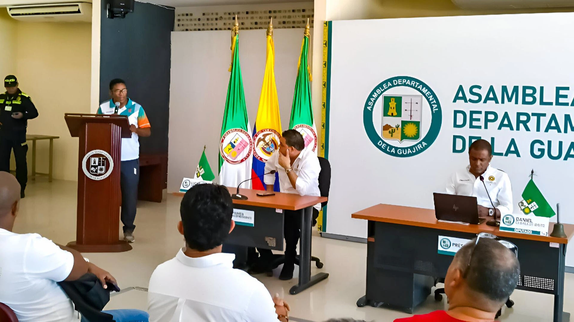 Funcionario expone en un atril durante una sesión en la Asamblea Departamental de La Guajira. Al fondo se ven banderas institucionales y dos miembros de la mesa directiva sentados frente a sus computadores, mientras los asistentes observan la intervención.