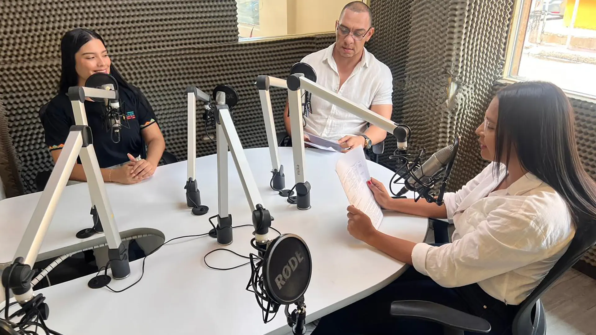 Tres personas se encuentran en una cabina de radio, sentadas alrededor de una mesa blanca equipada con micrófonos profesionales. Una mujer a la izquierda sonríe mientras conversa; frente a ella un hombre revisa unos documentos, y a la derecha una mujer sostiene hojas mientras habla al micrófono. El espacio está revestido con paneles acústicos y una ventana permite ver el exterior.