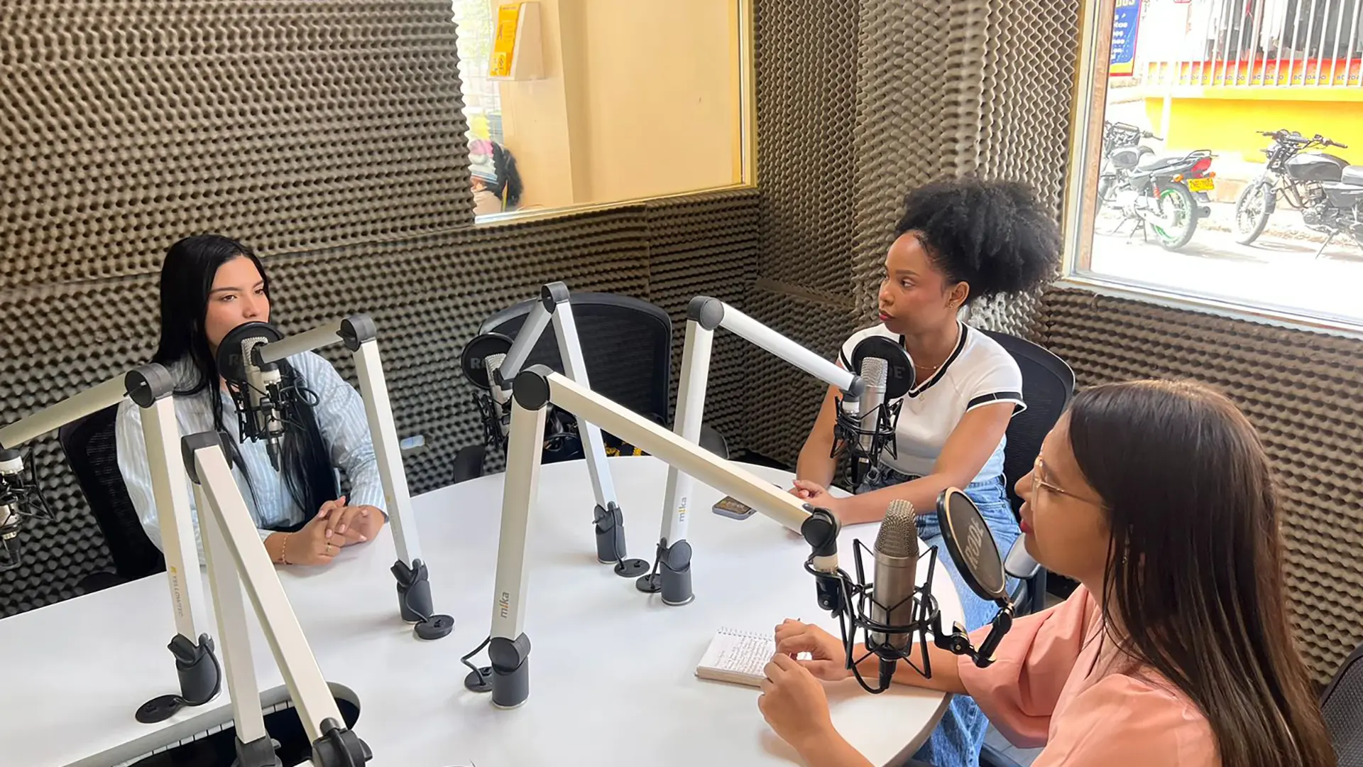 Tres mujeres conversan en una cabina de radio, sentadas alrededor de una mesa blanca equipada con micrófonos profesionales. Una de ellas toma notas en una libreta mientras las otras participan en el diálogo. El estudio está recubierto con paneles acústicos y cuenta con una ventana que muestra el exterior de la calle.