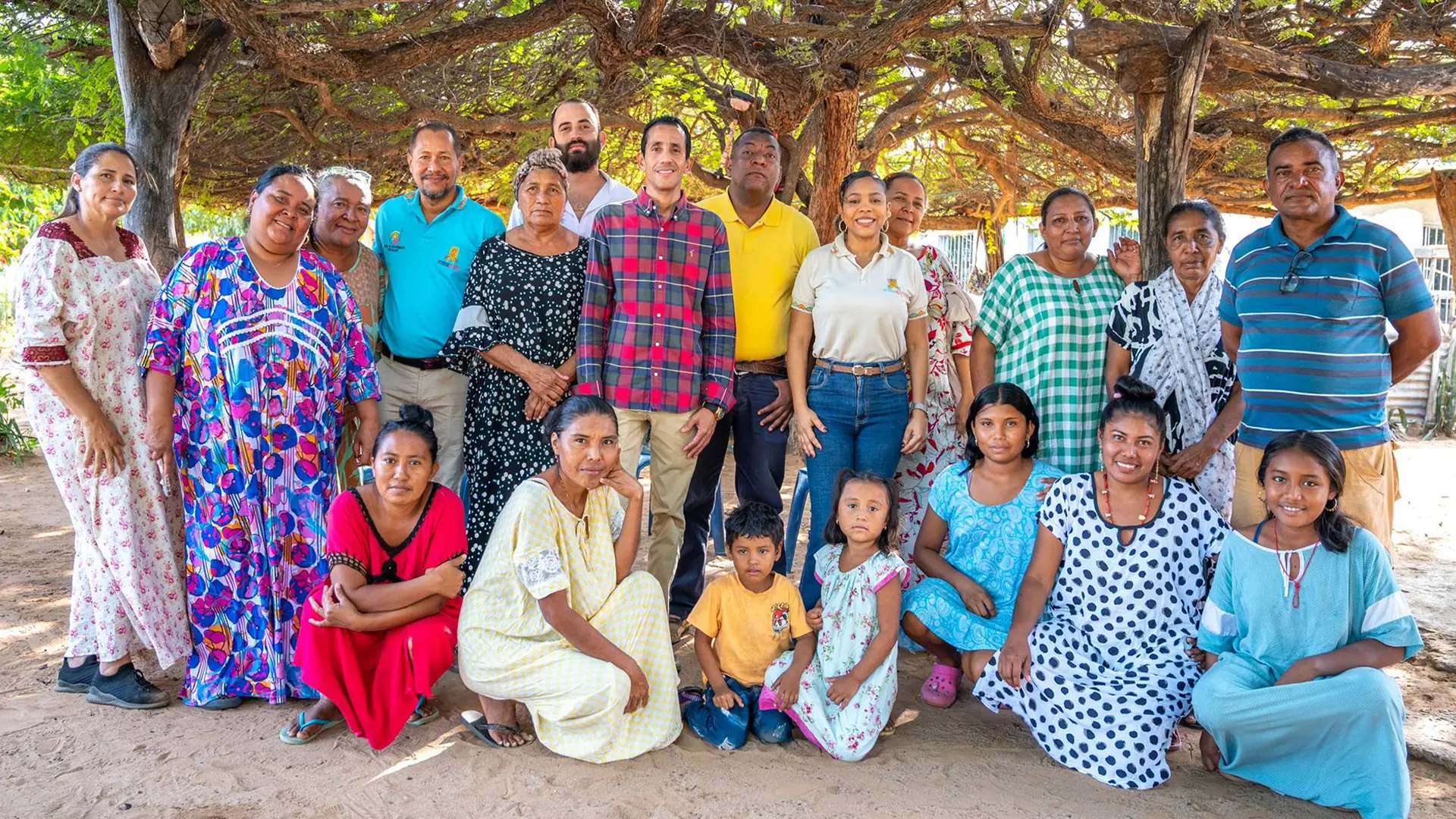 Grupo de personas adultas y niños posan juntos bajo un árbol en una comunidad rural, mostrando diversidad de edades y vestimenta, en un ambiente de encuentro comunitario.