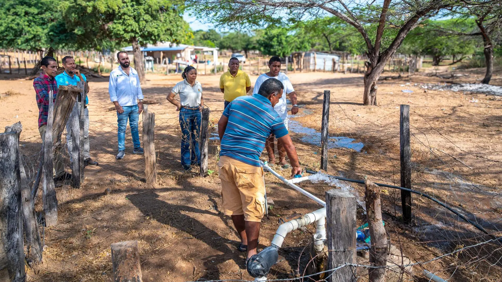 Grupo de personas inspecciona el funcionamiento de una tubería y válvula de agua instalada en una zona rural, rodeada de cercas de madera y árboles, como parte de una actividad comunitaria.