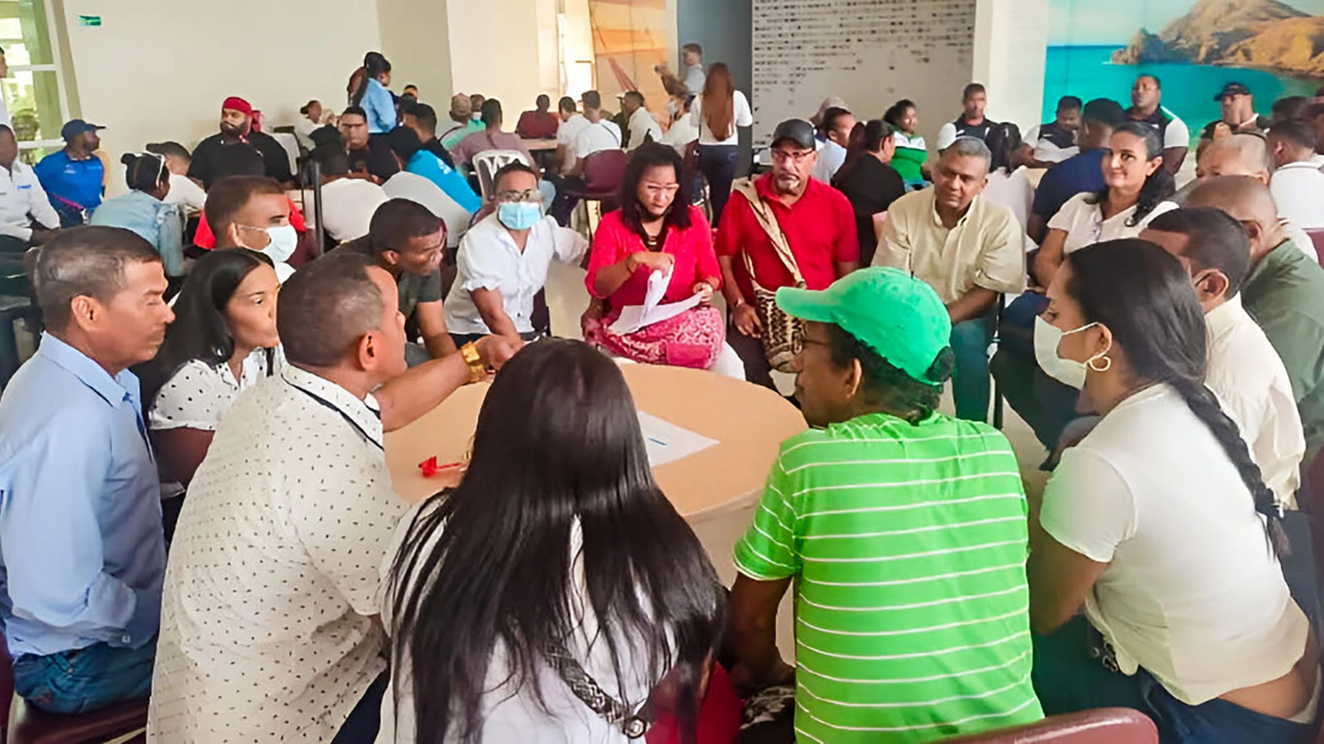 Grupo de participantes sentado alrededor de una mesa realizando una actividad de discusión en grupo, mientras otras personas conversan y trabajan en mesas al fondo en un salón amplio y bien iluminado.