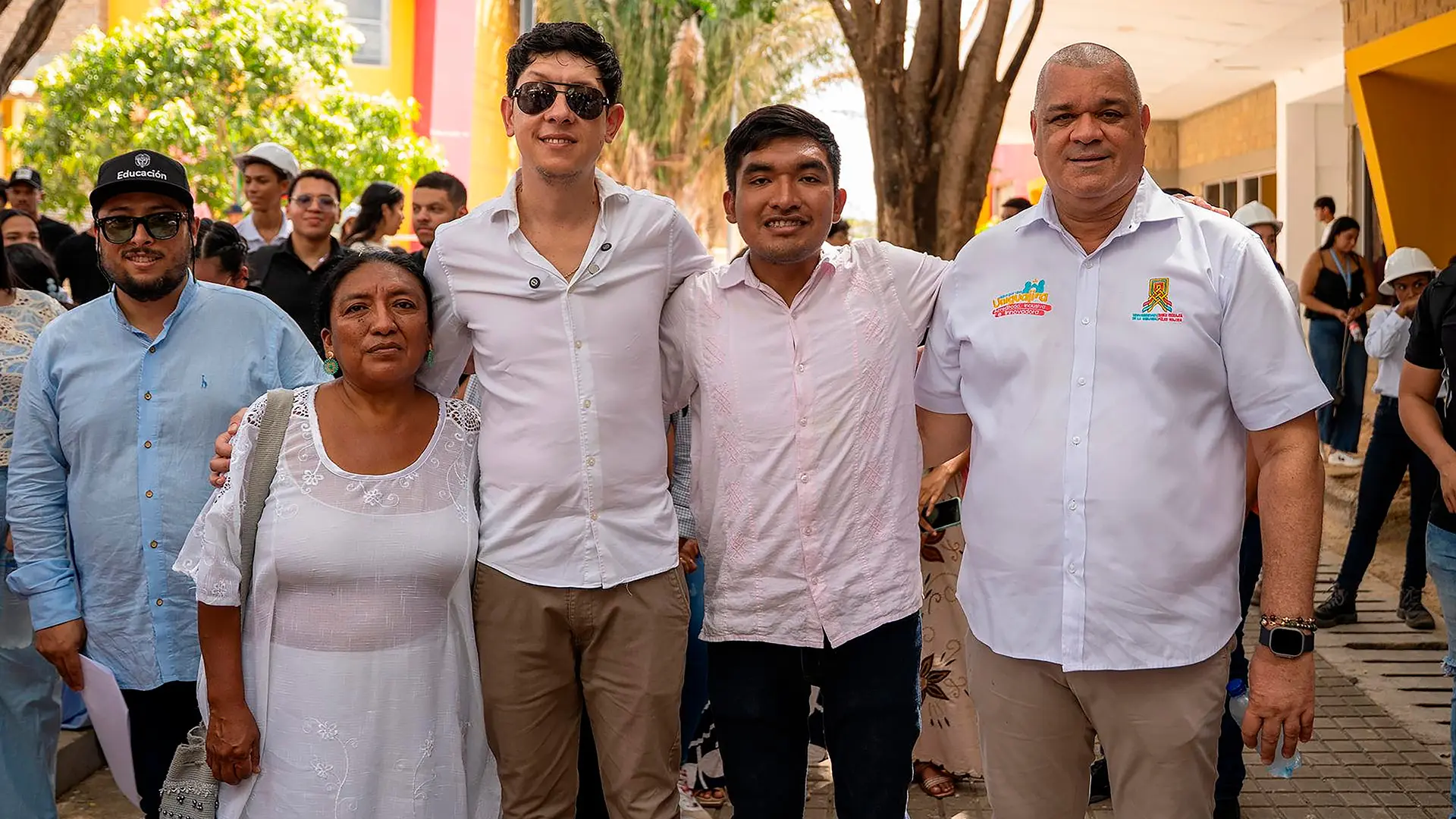 Cinco personas posan juntas durante una actividad pública al aire libre, rodeadas de asistentes. Dos hombres visten camisas blancas con logotipos institucionales, mientras el resto porta ropa casual y tradicional. El grupo aparece sonriente, con los brazos entrelazados, en un espacio arbolado frente a edificaciones coloridas, lo que sugiere un acto comunitario o institucional con amplia participación ciudadana.