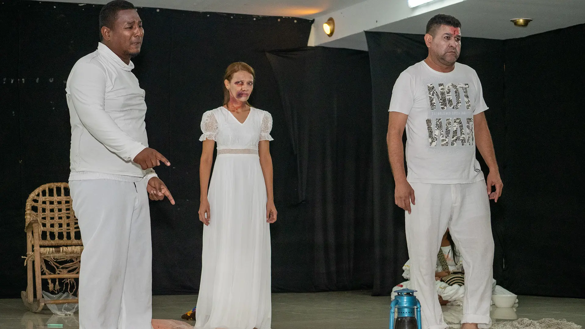 res actores sobre un escenario con fondo negro interpretan una escena teatral. Visten de blanco y muestran expresiones corporales intensas que transmiten tensión y dramatismo. En el centro, una mujer con vestido largo blanco permanece inmóvil, mientras dos hombres a los lados refuerzan la escena con gestos firmes. En el suelo se observan elementos escénicos que aportan simbolismo a la puesta en escena.