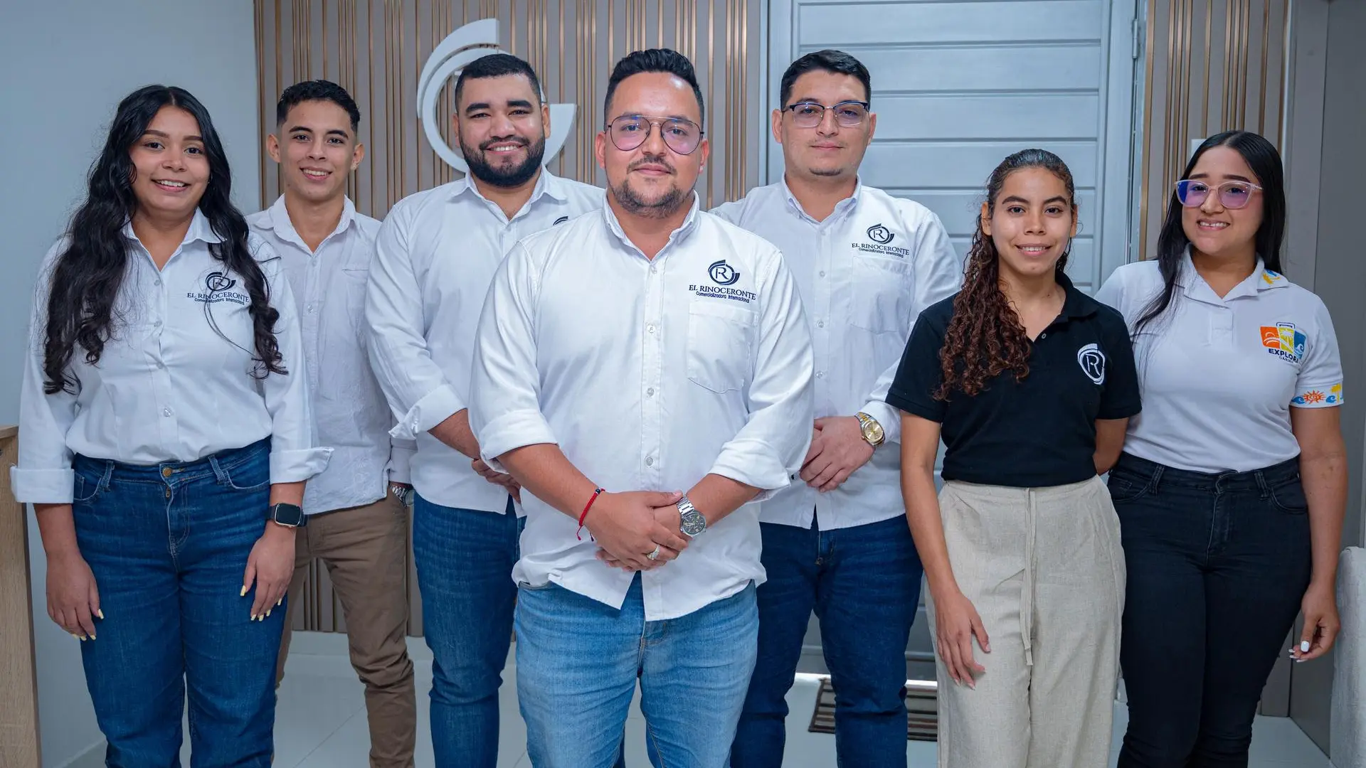 Siete integrantes de un equipo de trabajo posan en una oficina moderna, vestidos con camisas corporativas blancas y negras con logotipos institucionales, frente a una pared con diseño en madera y una puerta al fondo.