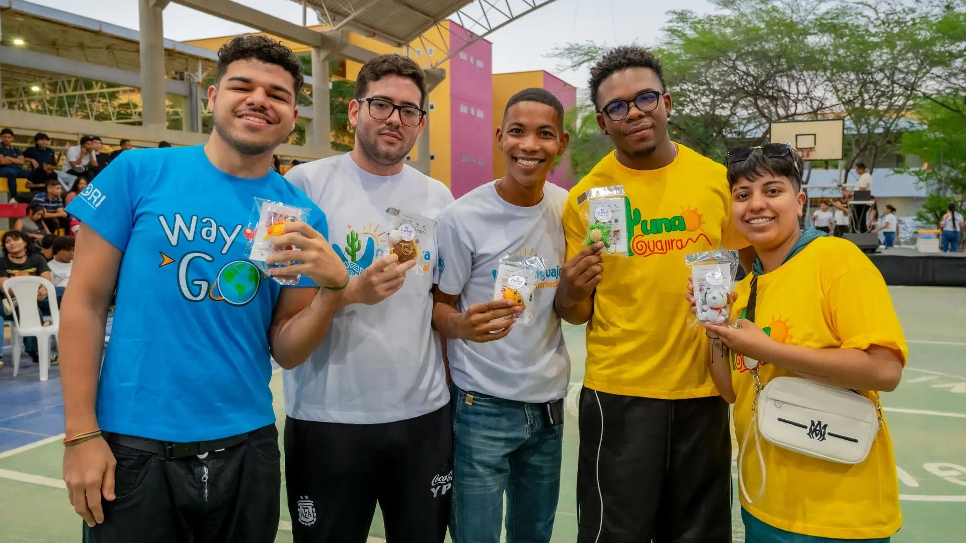 Grupo de seis jóvenes sonriendo en una cancha deportiva durante un evento universitario, sosteniendo pequeños paquetes promocionales. Algunos visten camisetas institucionales y el fondo muestra estudiantes sentados y estructuras del campus.