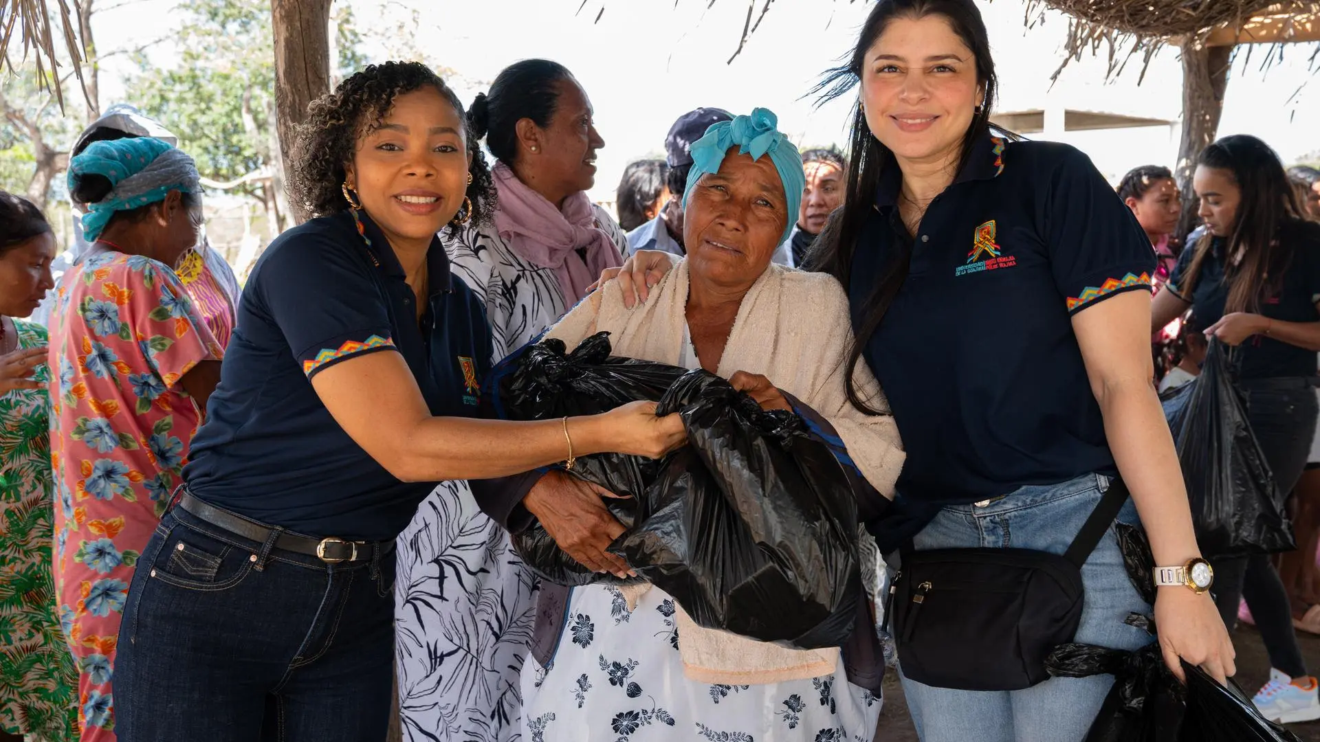 Dos mujeres entregan bolsas de ayuda humanitaria a una adulta mayor en una comunidad rural, rodeadas de otras personas en un entorno comunitario. La escena representa solidaridad, apoyo social, asistencia humanitaria y trabajo comunitario.