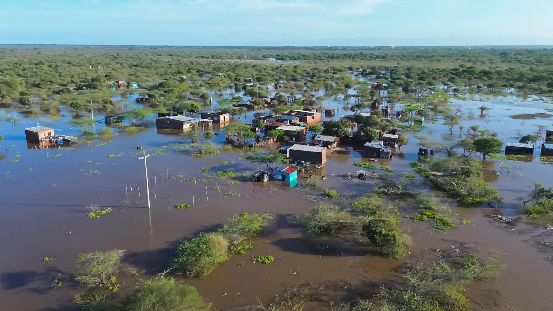 Vista aérea de un asentamiento rural severamente inundado en una zona de vegetación baja y arbustiva. Las viviendas, construidas con materiales sencillos, están rodeadas por agua turbia que cubre los caminos y patios. Se observan postes de luz sumergidos en el agua y árboles emergiendo de la inundación bajo un cielo despejado. La imagen captura la magnitud de un desastre natural en una comunidad vulnerable.