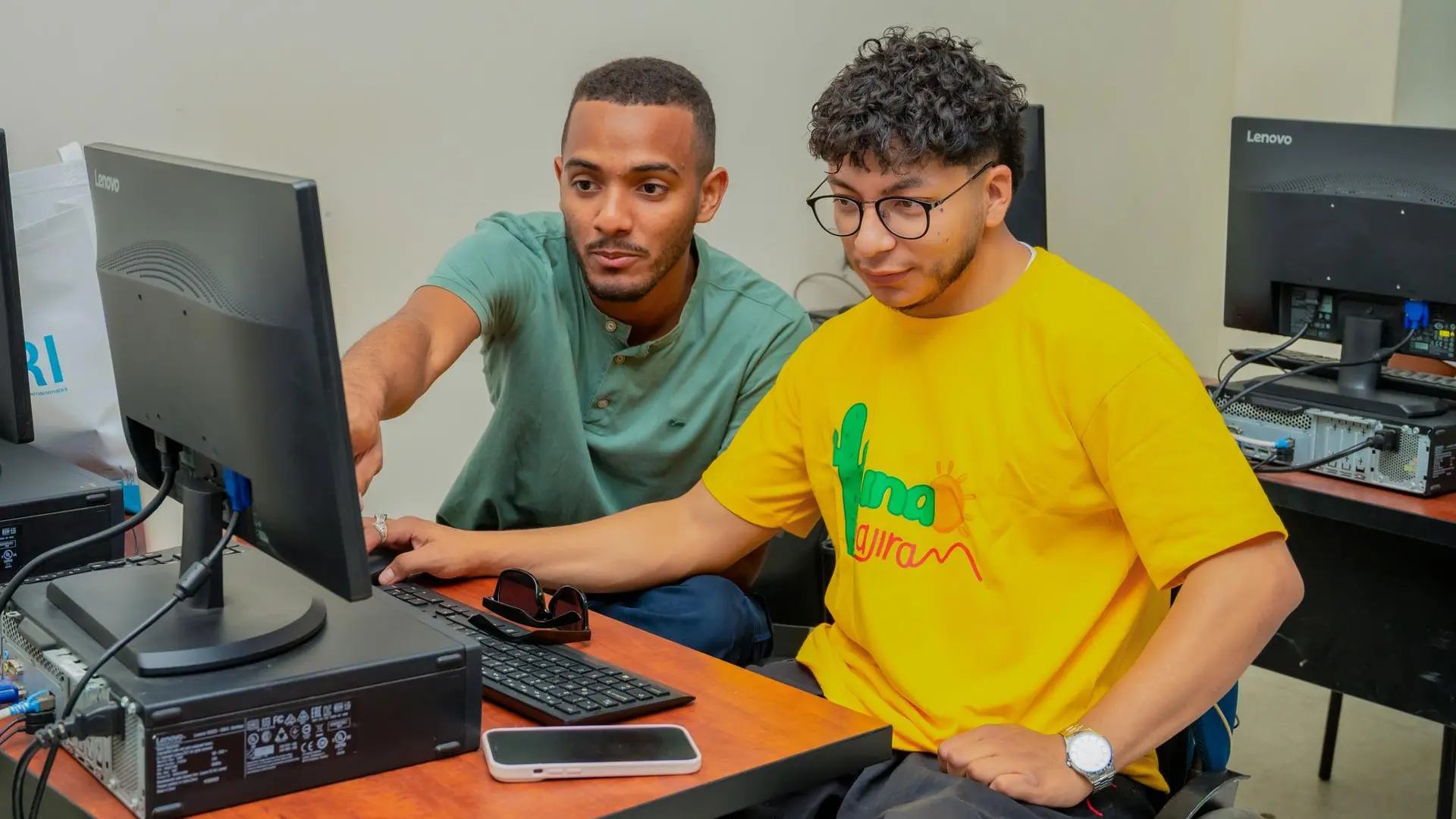 Dos jóvenes trabajan juntos frente a un computador en una sala de informática; uno señala la pantalla mientras el otro usa el teclado. Ambos están concentrados en la actividad, en un ambiente académico con equipos de escritorio alrededor.”