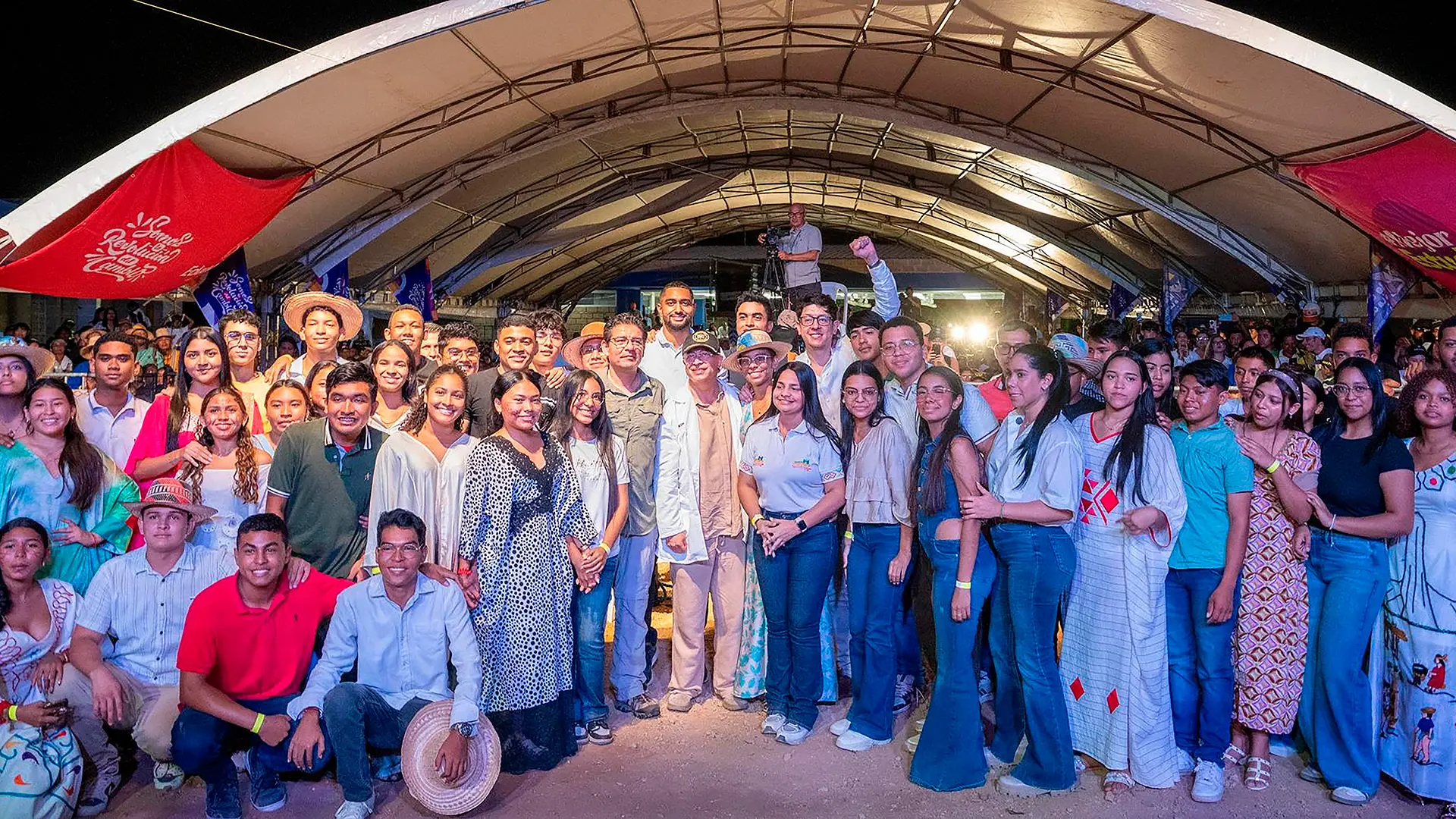 Un numeroso grupo de jóvenes y adultos posa para una fotografía colectiva durante un evento cultural realizado de noche, bajo una gran carpa iluminada. Los asistentes, vestidos con ropa cotidiana y prendas tradicionales, sonríen y levantan los brazos en señal de entusiasmo, mientras banderas y pancartas rodean el espacio. Al fondo se observa público, personal técnico y luces de escenario, lo que refuerza el carácter festivo y comunitario del encuentro.