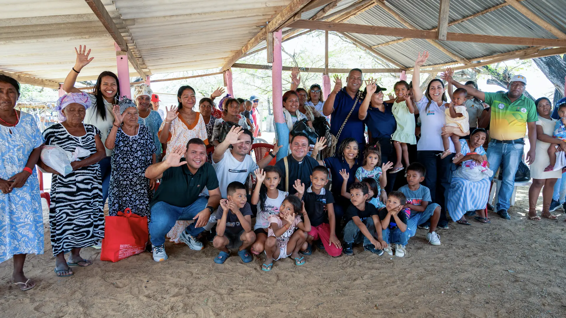 Grupo grande de personas de una comunidad rural, incluyendo niños, jóvenes, adultos y adultos mayores, reunidos bajo una estructura con techo de zinc, saludando y sonriendo a la cámara. La escena refleja unión comunitaria, participación social y convivencia intergeneracional.