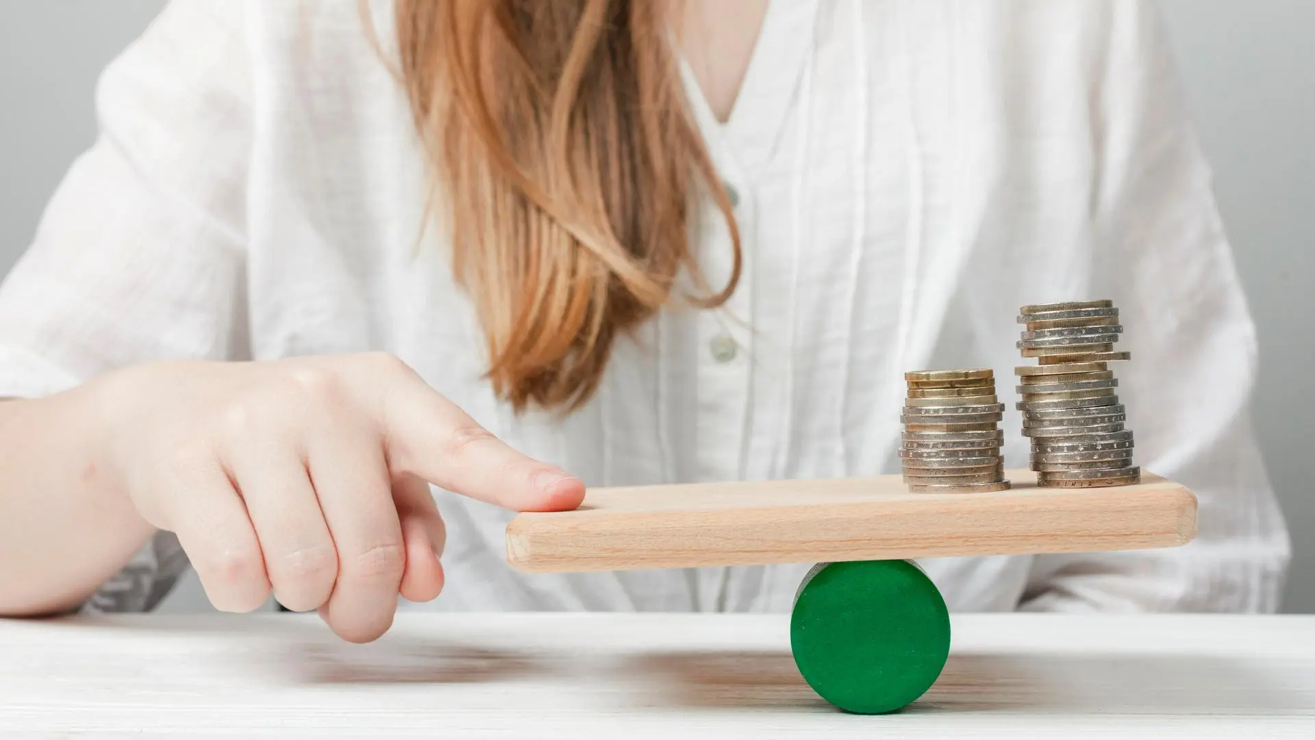 Mano de una mujer equilibrando una tabla de madera sobre un cilindro verde, con dos montones de monedas apiladas que representan un balance económico.
