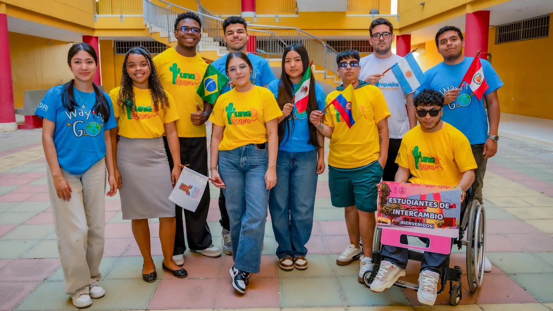 Grupo diverso de estudiantes universitarios en un patio institucional, vestidos con camisetas de colores representando un programa internacional. Algunos sostienen banderas de diferentes países y un estudiante en silla de ruedas sostiene un cartel de bienvenida para estudiantes de intercambio.