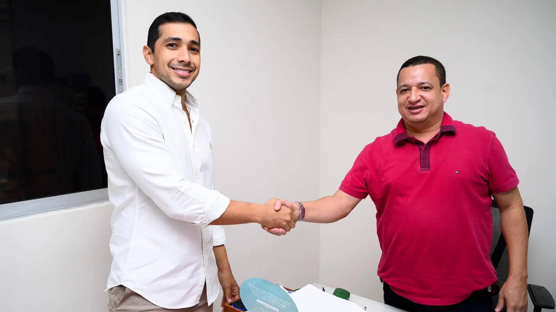 Dos hombres estrechan la mano en una oficina, uno vestido con camisa blanca y el otro con camiseta roja. Están junto a un escritorio en un ambiente interior iluminado. La escena transmite un saludo cordial entre ambos.