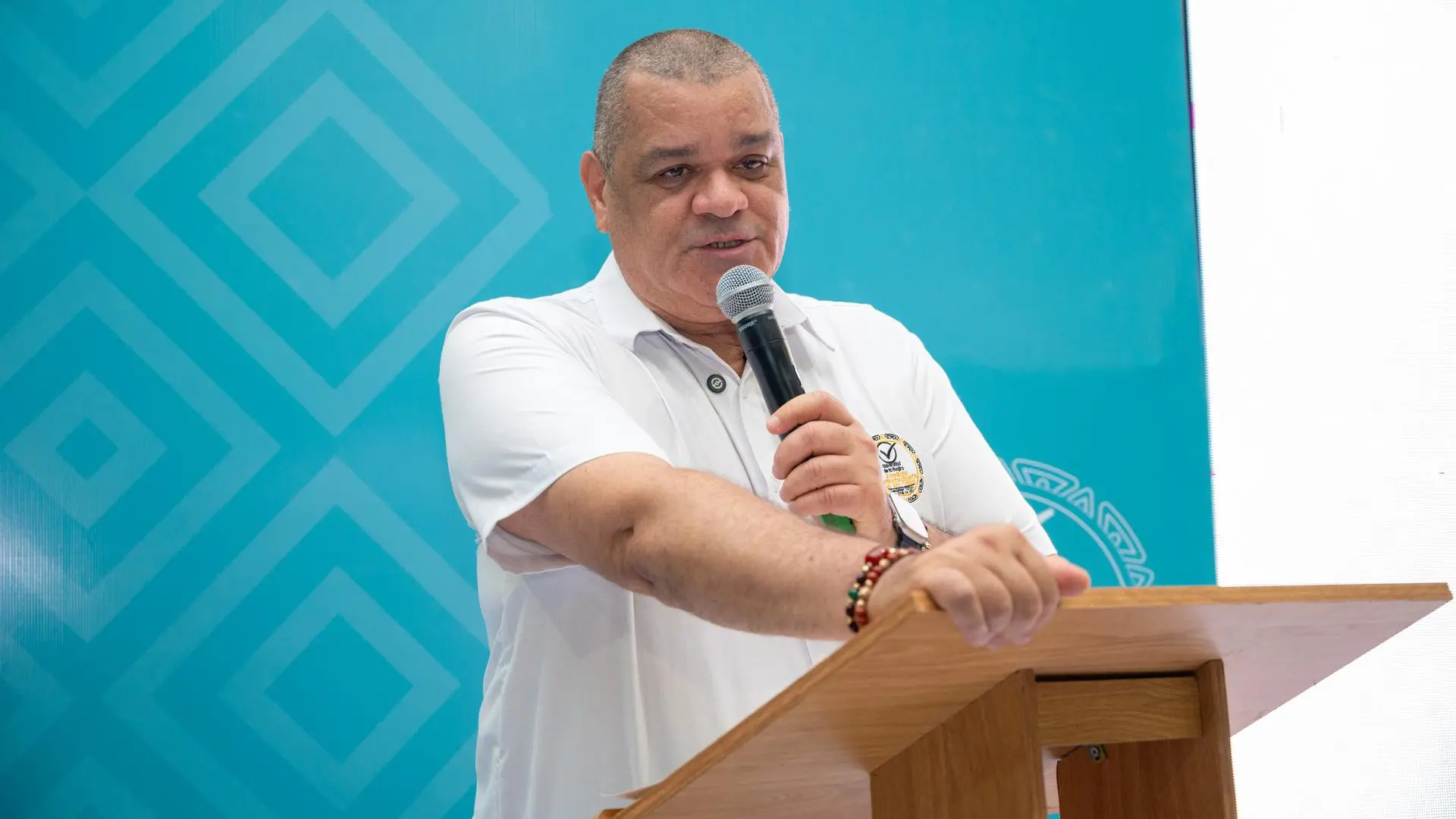 Un hombre sostiene un micrófono mientras habla desde un atril con expresión seria y concentrada. Viste una camiseta blanca y se encuentra frente a un fondo azul con patrones geométricos. La escena sugiere una presentación o discurso en un entorno formal.