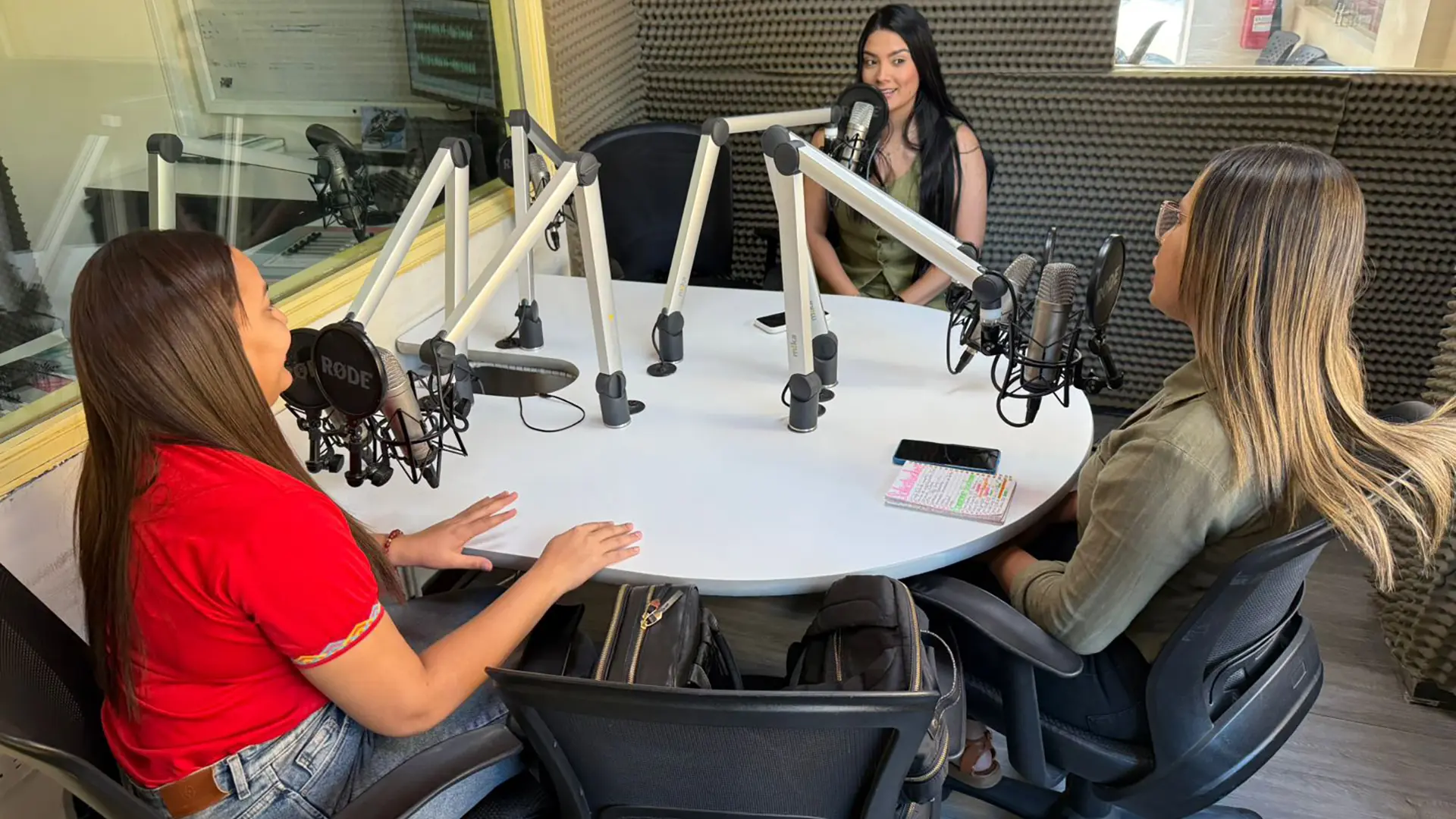 Tres mujeres participan en una conversación dentro de una cabina de radio, sentadas alrededor de una mesa con micrófonos profesionales. Una de ellas habla mientras las otras escuchan y toman notas. El estudio está recubierto con paneles acústicos y tiene una ventana hacia una sala de control.