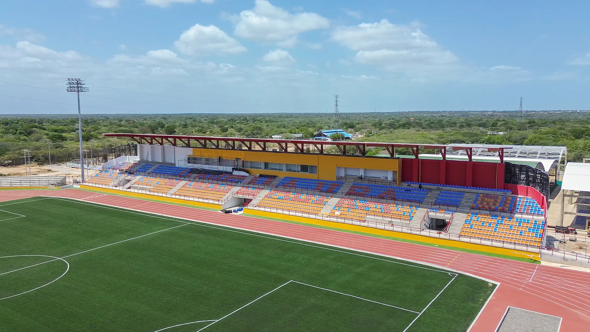 Vista aérea de un estadio con cancha de fútbol y pista atlética, graderías multicolores techadas y entorno natural alrededor.