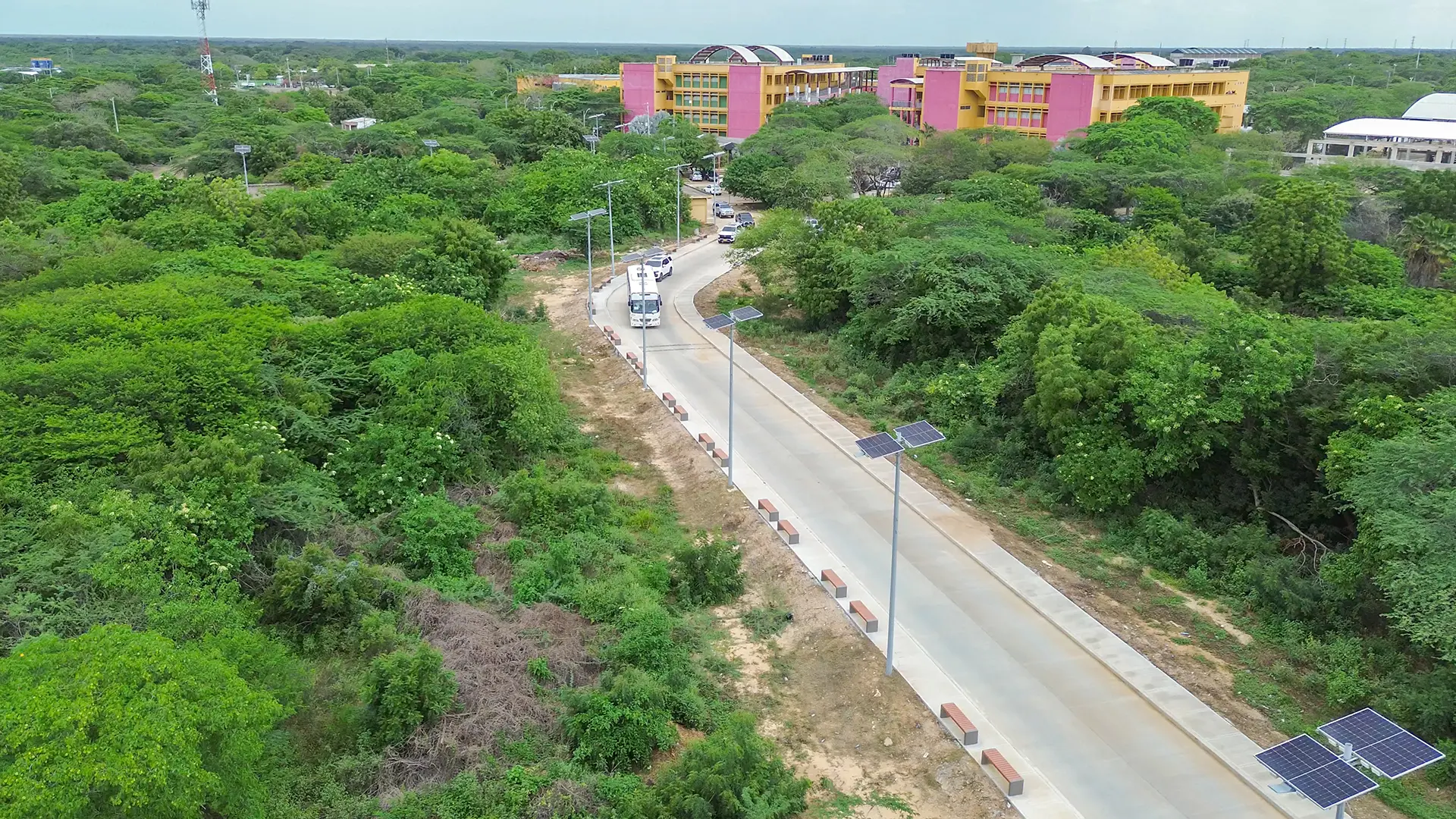 Vista aérea de una vía pavimentada rodeada de vegetación densa, con postes de alumbrado con paneles solares y vehículos en circulación, que conduce hacia edificaciones académicas de colores.