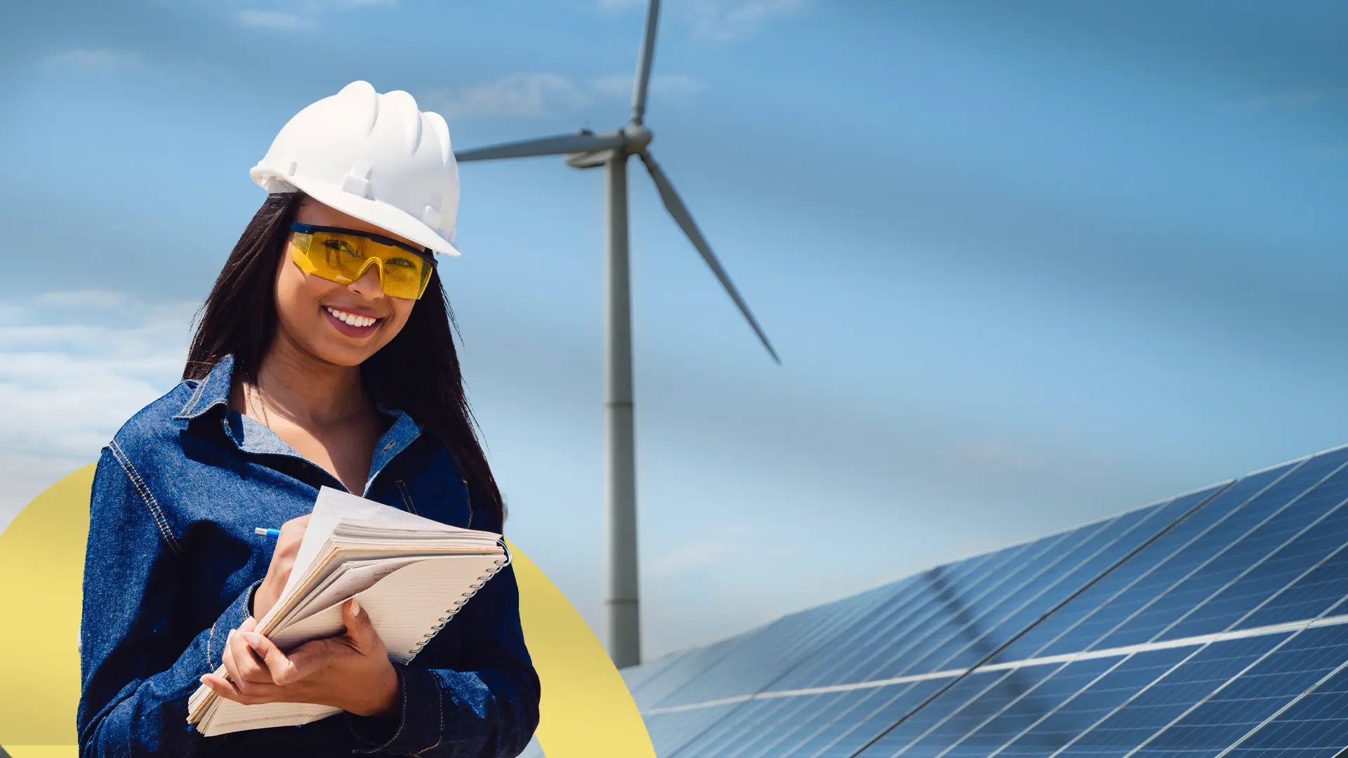 Persona con casco de seguridad y gafas protectoras sostiene una libreta mientras sonríe, aparentemente tomando notas. Al fondo se observan paneles solares y un aerogenerador, en un entorno de energía renovable bajo cielo despejado. La escena transmite trabajo técnico y sostenibilidad energética.