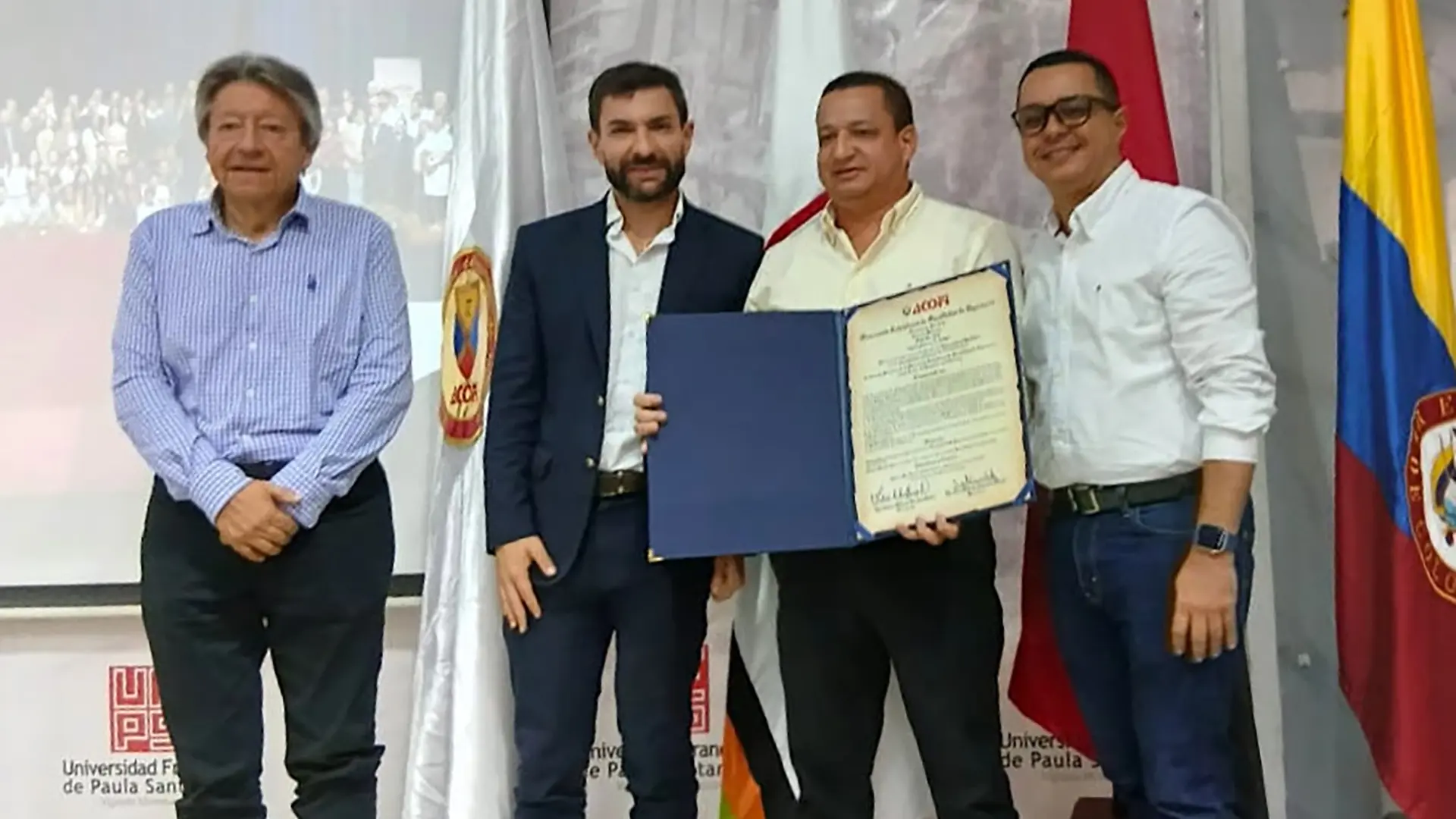 En un auditorio, cuatro hombres posan de pie frente a un fondo institucional de la Universidad Francisco de Paula Santander y varias banderas, entre ellas la de Colombia. En el centro, uno de ellos sostiene un diploma o reconocimiento abierto mientras los demás lo acompañan a su lado. La escena corresponde a una ceremonia de reconocimiento, con una pantalla de proyección visible al fondo.