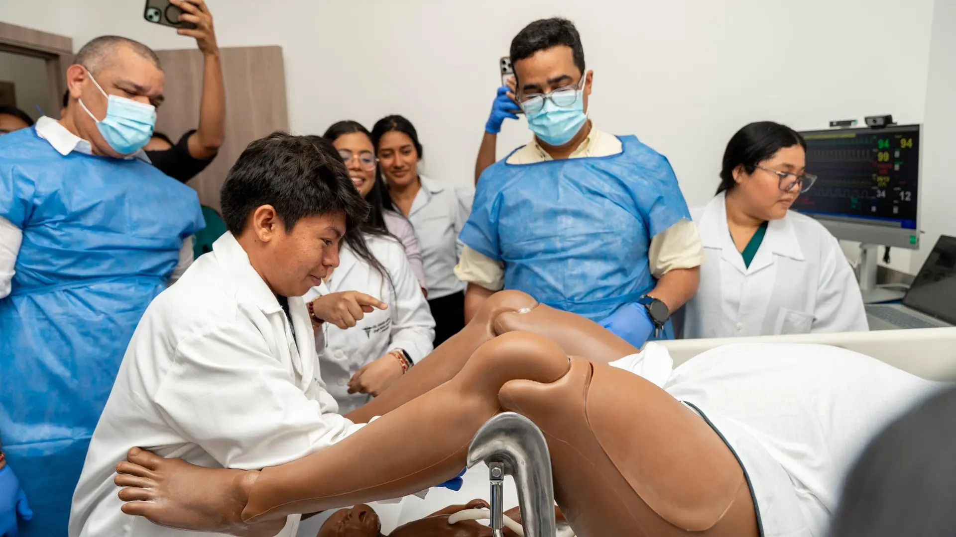 Grupo de estudiantes de salud realizando una práctica clínica con un maniquí de simulación, mientras un instructor supervisa el procedimiento en un entorno médico equipado.