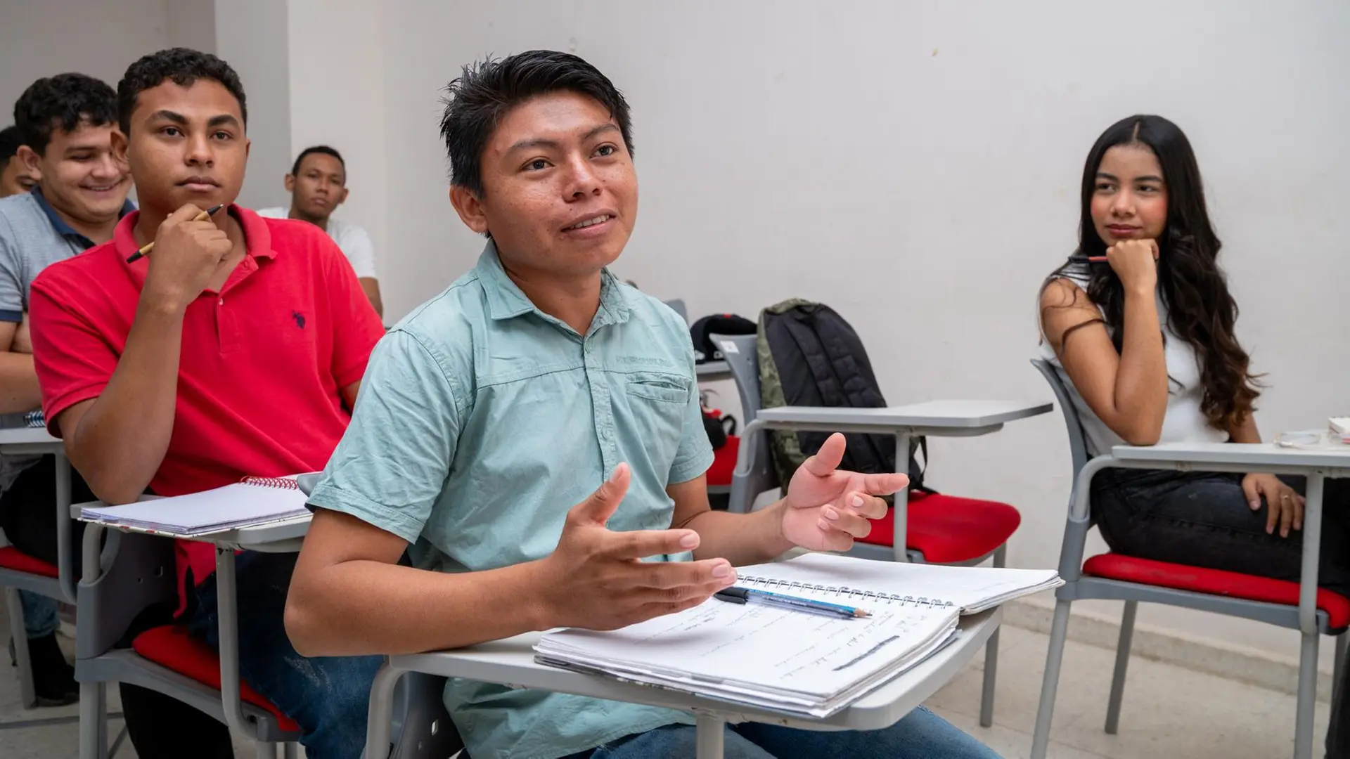 Estudiantes sentados en un salón de clases, uno de ellos participa activamente mientras habla y gesticula con las manos, mientras sus compañeros escuchan y toman apuntes.