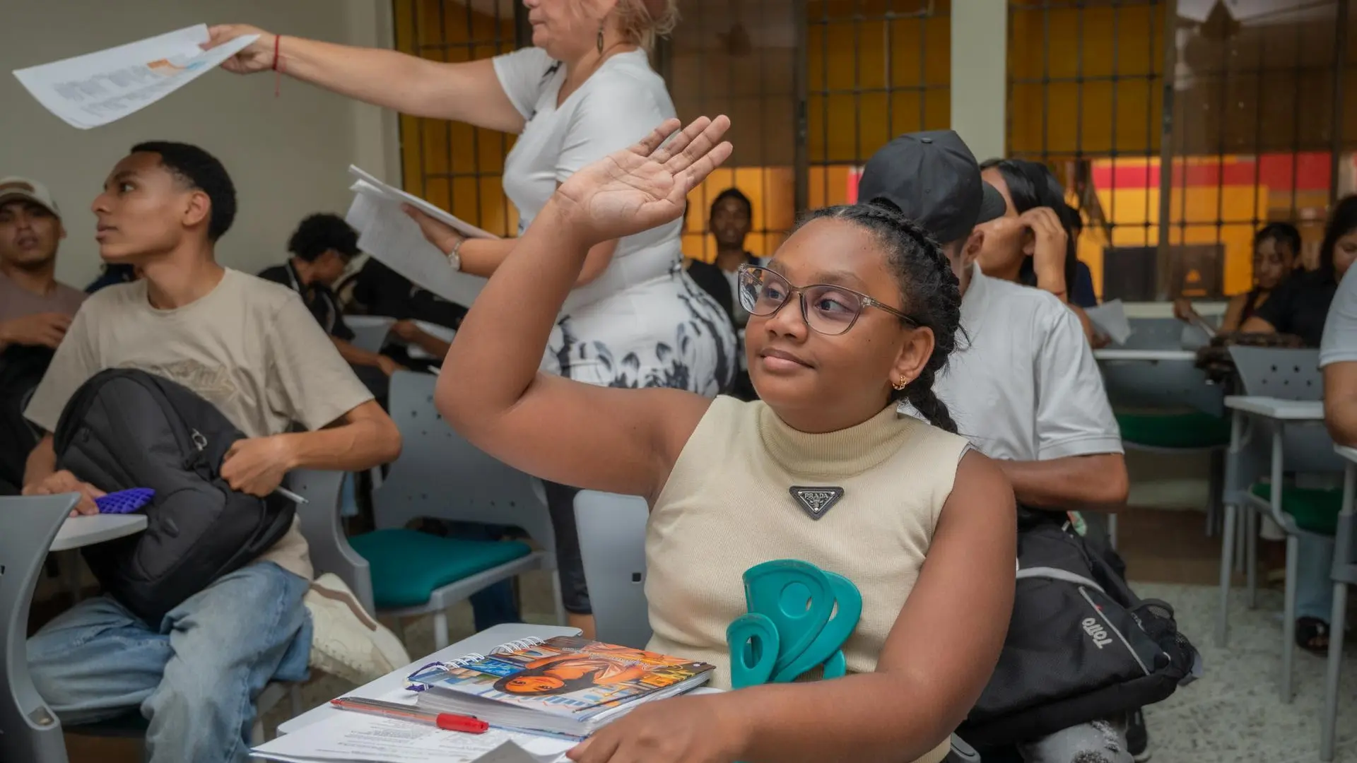 Estudiante levanta la mano en un aula mientras sostiene una muleta y cuadernos en su escritorio.