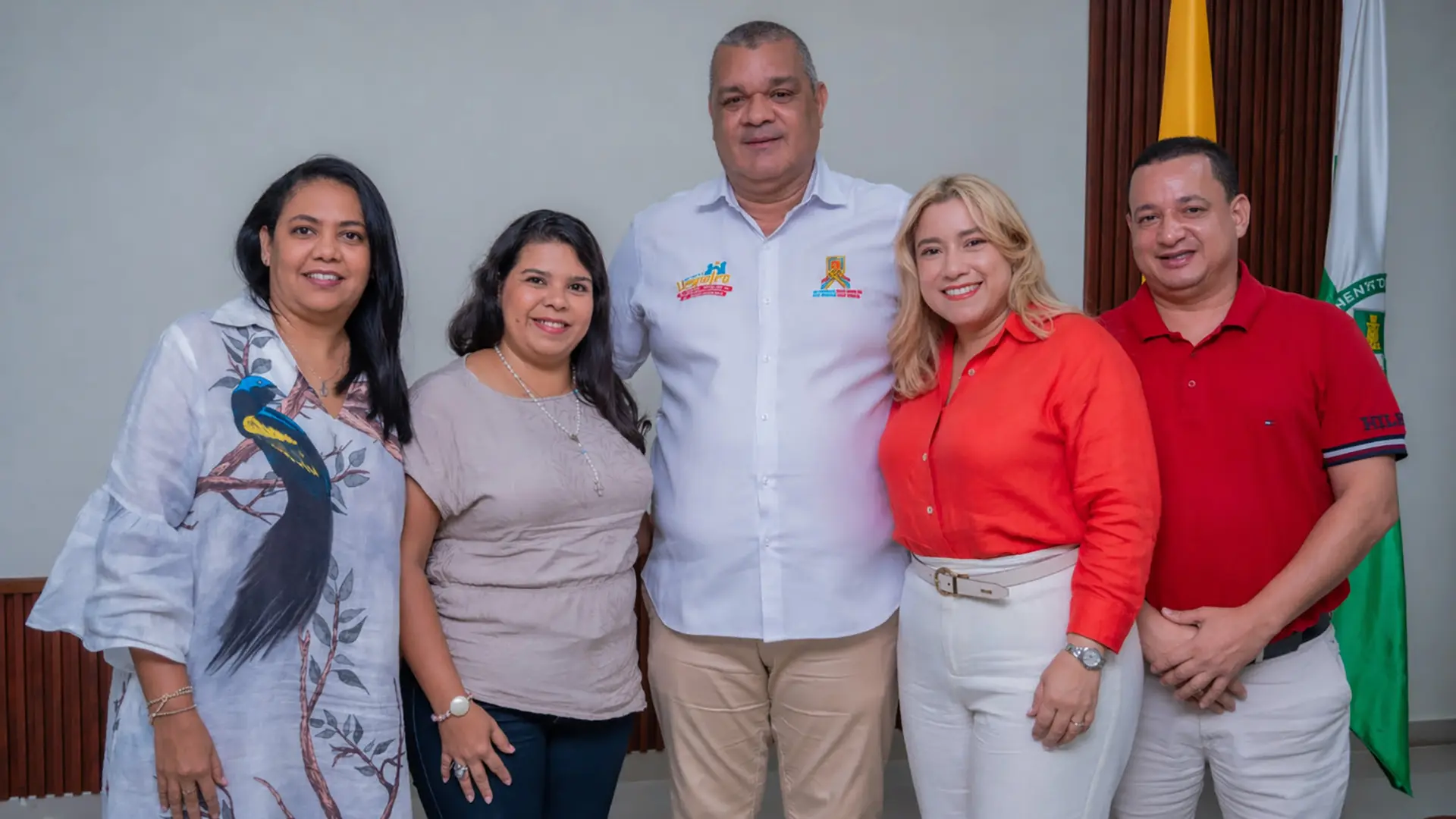 Un grupo de cinco personas, tres mujeres y dos hombres, posan sonrientes para un retrato en un espacio interior. Los asistentes visten de forma casual-formal, destacando una mujer con una blusa naranja vibrante y un hombre en el centro con una camisa blanca que lleva logotipos bordados. A la derecha de la imagen, se observan parcialmente dos banderas que sugieren un contexto institucional o gubernamental.