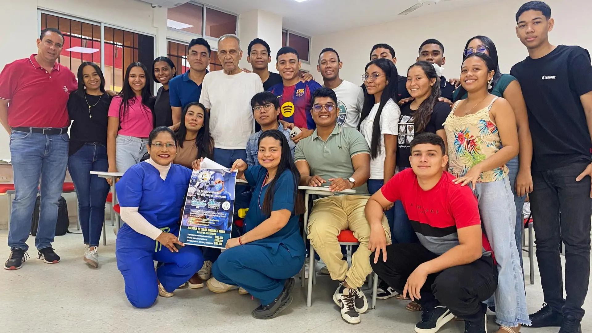Grupo de estudiantes y docentes posan en un aula, sonriendo a la cámara. En el centro, un hombre mayor se encuentra rodeado por los jóvenes, mientras dos mujeres en uniforme de salud, ubicadas al frente, sostienen un afiche informativo. El salón cuenta con escritorios, sillas, aire acondicionado y ventilador en el techo. La escena refleja un ambiente académico y colaborativo, posiblemente relacionado con una actividad formativa o institucional.