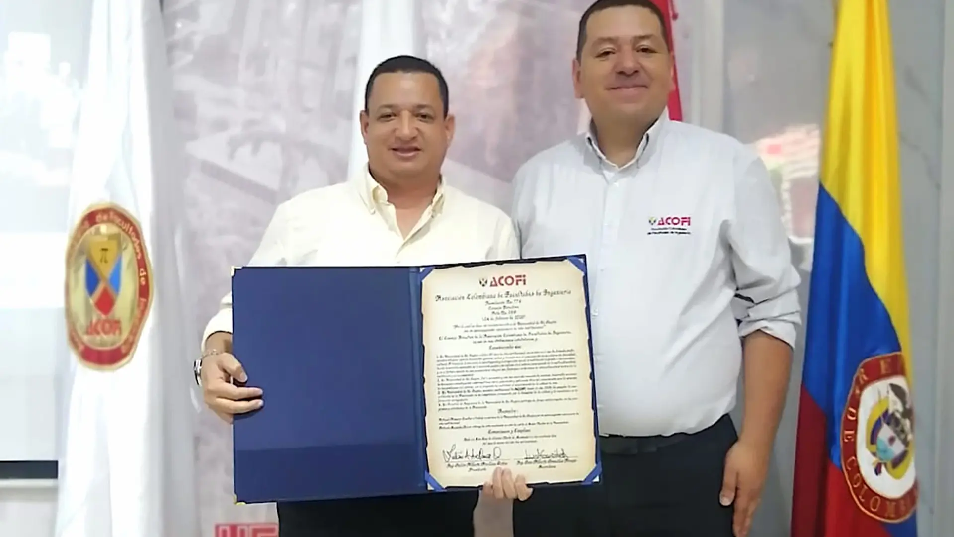 Dos hombres posan de pie en un acto institucional mientras sostienen un reconocimiento abierto otorgado por ACOFI. Detrás de ellos se observa un fondo con el logotipo de la Universidad Francisco de Paula Santander y varias banderas institucionales y la bandera de Colombia. La escena corresponde a un acto de reconocimiento realizado en un espacio universitario.