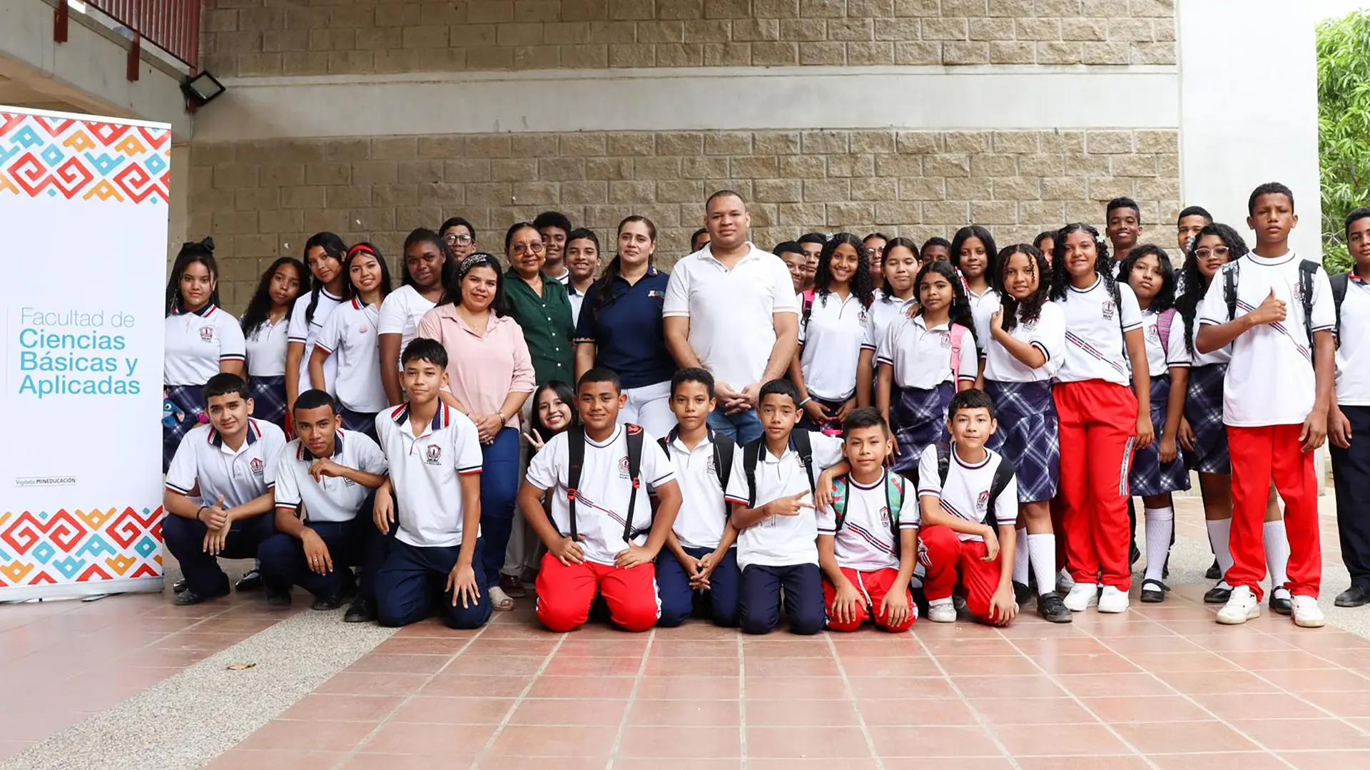Grupo grande de estudiantes de primaria y docentes posando juntos en un patio escolar. Visten uniformes blancos con detalles rojos y azules. A un lado hay un banner institucional de la Facultad de Ciencias Básicas y Aplicadas. El entorno es un espacio abierto con paredes de ladrillo y piso de baldosas.