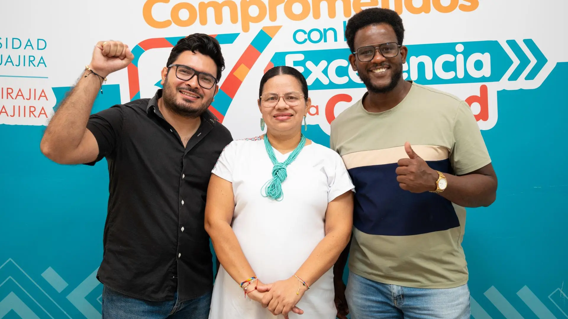 Tres personas posan sonrientes frente a un fondo institucional con mensajes sobre excelencia; uno levanta el puño en señal de entusiasmo mientras otro hace un gesto de aprobación con el pulgar.