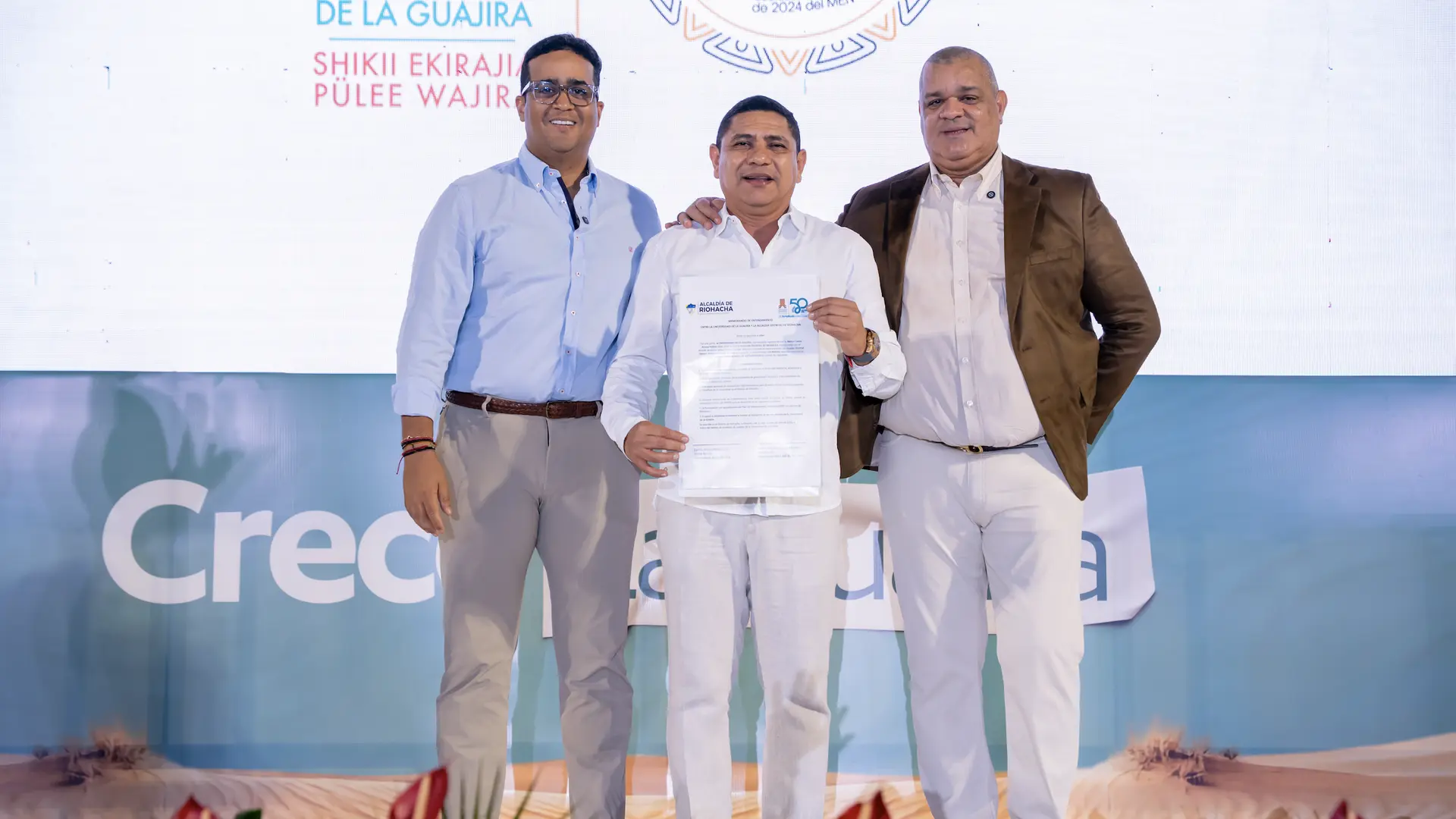 Tres hombres posan sobre un escenario institucional frente a un fondo con el nombre de la Universidad de La Guajira. El hombre del centro sostiene un documento oficial mientras los otros dos lo acompañan con gestos de apoyo, en un ambiente formal de ceremonia o acto de reconocimiento.