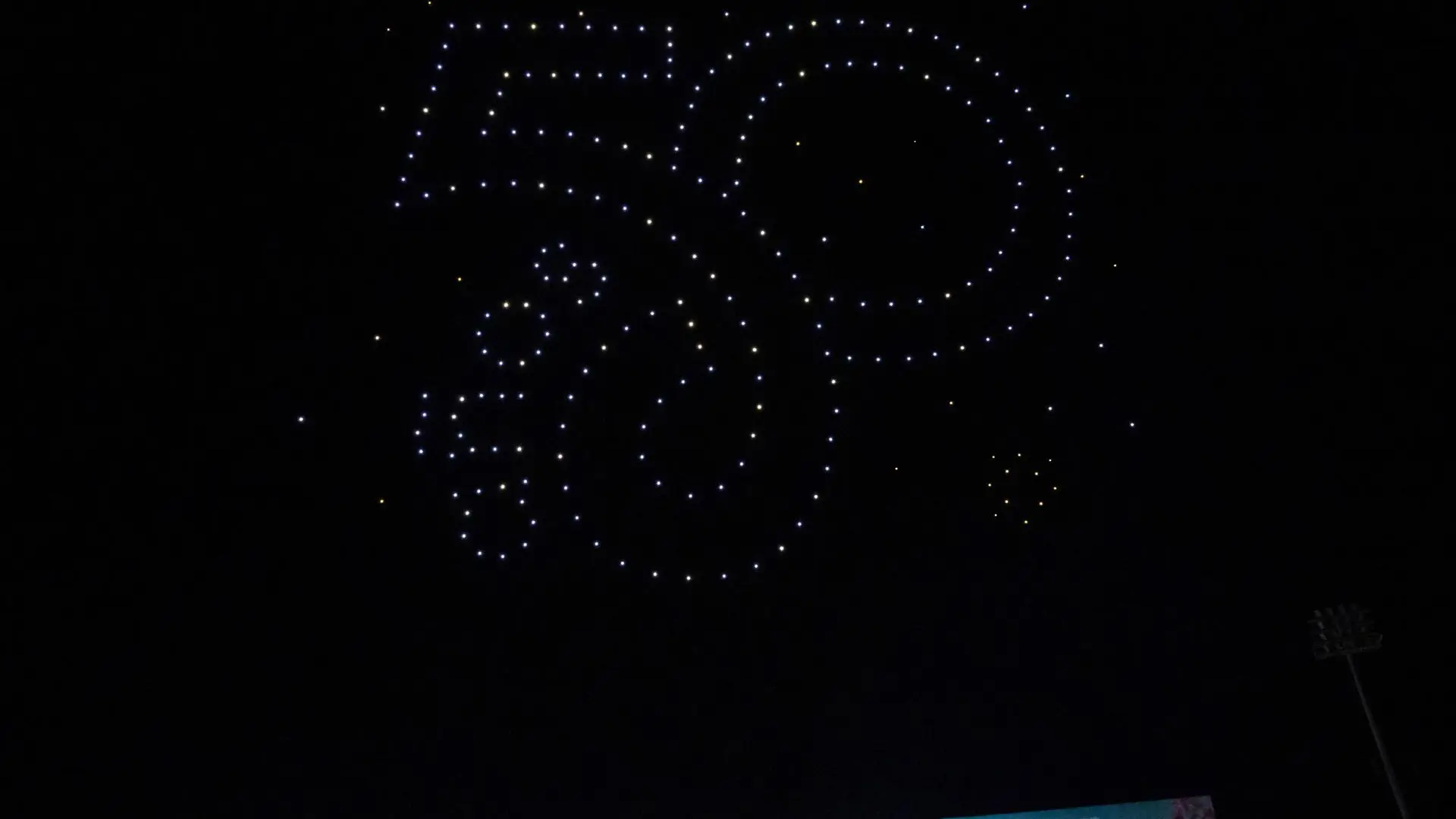 Espectáculo nocturno de drones que forman en el cielo la figura del número “50” junto a un diseño decorativo, compuesto por múltiples luces blancas y amarillas. En la parte inferior se alcanza a ver la tarima del evento, sugiriendo una celebración conmemorativa al aire libre.