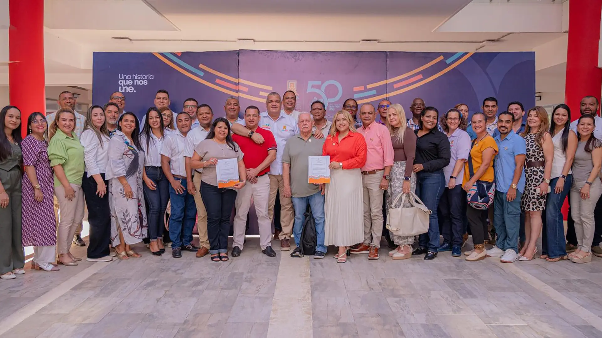 Fotografía grupal muestra a unas cuarenta personas posando En el centro, un hombre y una mujer sostienen certificados oficiales, todos visten ropa casual y formal en tonos variados. La imagen captura un ambiente de celebración institucional, con los asistentes sonriendo de manera organizada para conmemorar el aniversario.