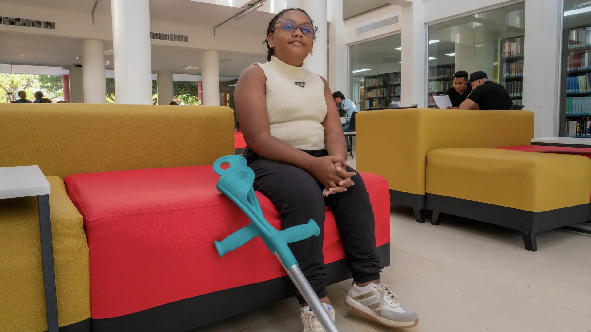 Persona joven sentada en un sillón rojo dentro de una biblioteca, con muleta apoyada a su lado.