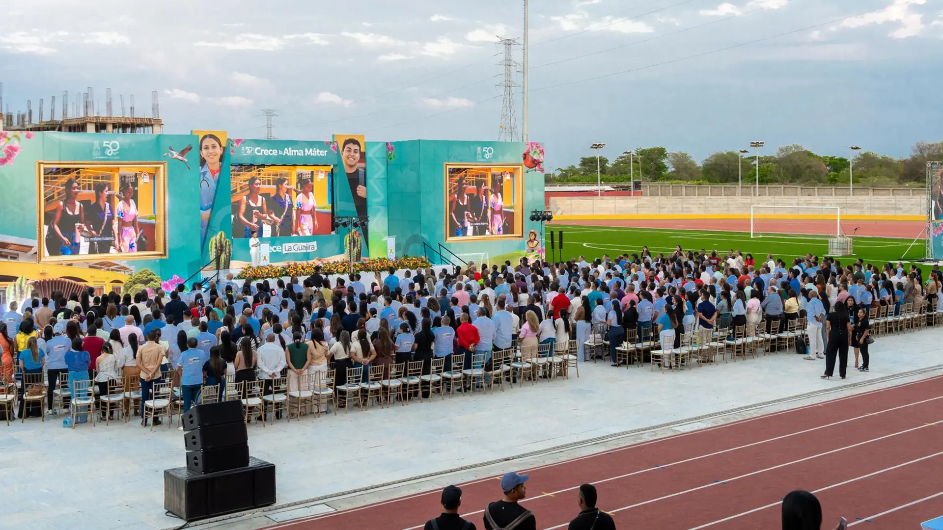 Multitud de personas asiste a un evento institucional al aire libre en una cancha deportiva. Frente a ellas, un escenario amplio y colorido con pantallas gigantes proyecta imágenes de estudiantes y mensajes educativos. El público, de pie junto a filas de sillas, observa la presentación en un ambiente organizado y masivo, con cielo parcialmente nublado al fondo.