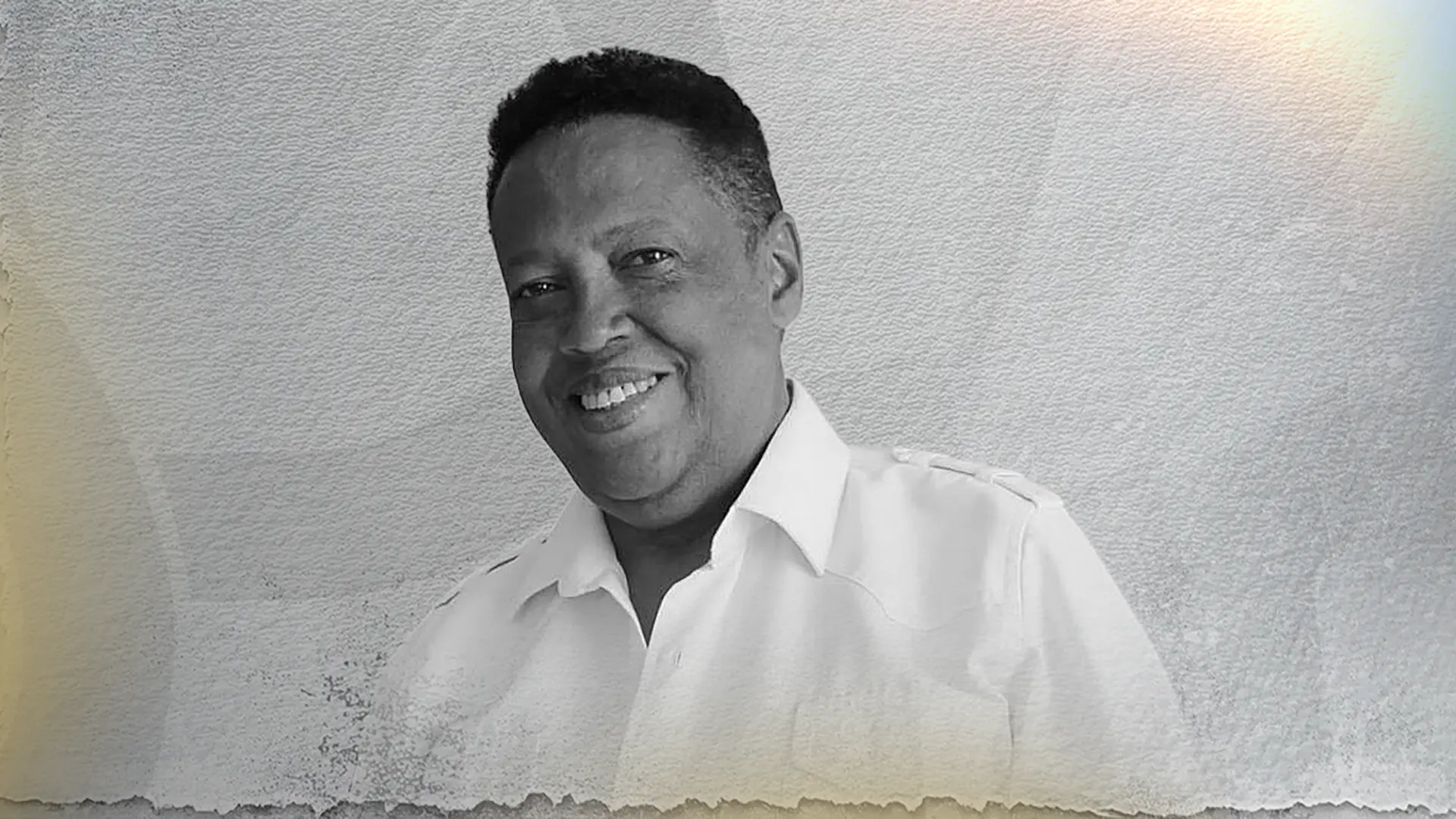Fotografía en blanco y negro de un hombre sonriente posando frente a una pared clara con textura. Lleva una camisa blanca de botones y mira directamente a la cámara con una expresión amable y relajada. La imagen tiene un efecto artístico con bordes desgastados y una iluminación suave que aporta un estilo elegante y nostálgico.