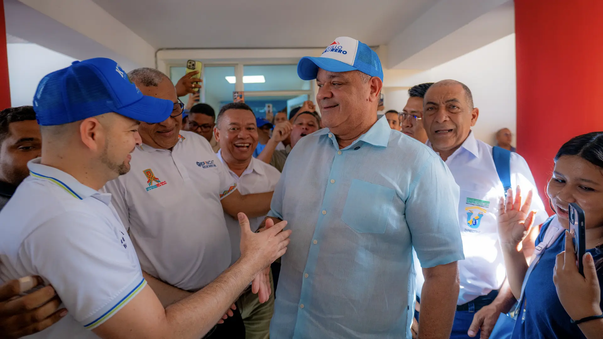 Un grupo de personas recibe cordialmente a un líder institucional en un espacio interior iluminado. La escena refleja cercanía, diálogo y apoyo comunitario, mientras varios asistentes sonríen y registran el momento con sus celulares. Predominan tonos azules y blancos en un ambiente dinámico y participativo.