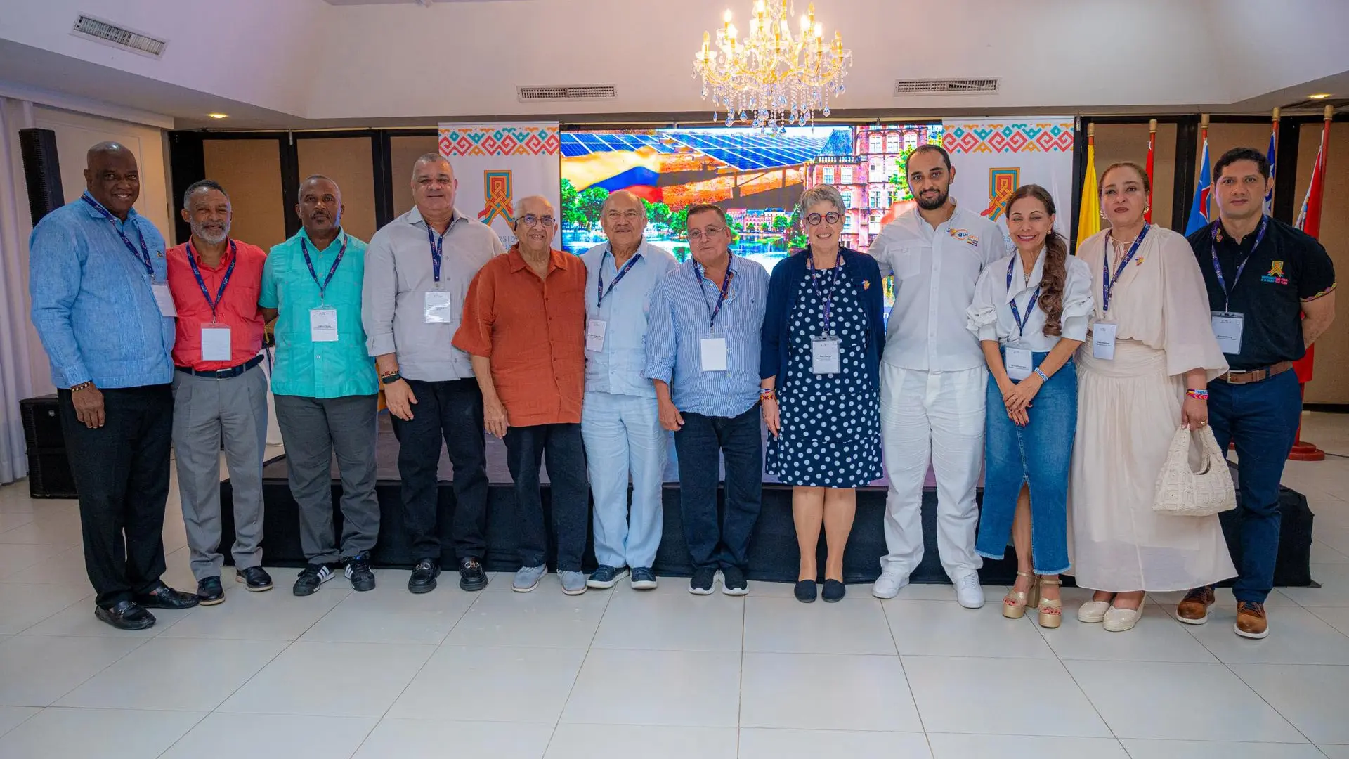 Un grupo de once personas posa alineado en un salón de eventos bajo una elegante lámpara de cristal para una fotografía oficial. Los asistentes visten ropa casual elegante y portan credenciales del evento frente a una pantalla con imágenes de paneles solares y banderas. El entorno sugiere una clausura o encuentro académico en un auditorio.