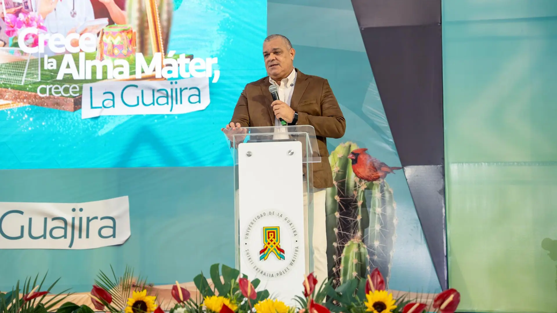Un hombre con chaqueta marrón pronunciando un discurso detrás de un podio con el logotipo de la Universidad de La Guajira en un evento. Detrás de él, una gran pantalla muestra el eslogan "Crece la Alma Máter, Crece La Guajira" y fotos de estudiantes.