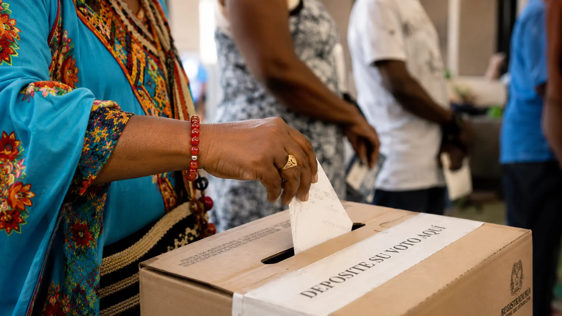 Primer plano de una persona depositando su voto en una urna electoral de la Registraduría Nacional. La imagen destaca la mano de la votante y su vestimenta colorida con detalles artesanales, mientras otras personas esperan al fondo en un espacio de votación interior.
