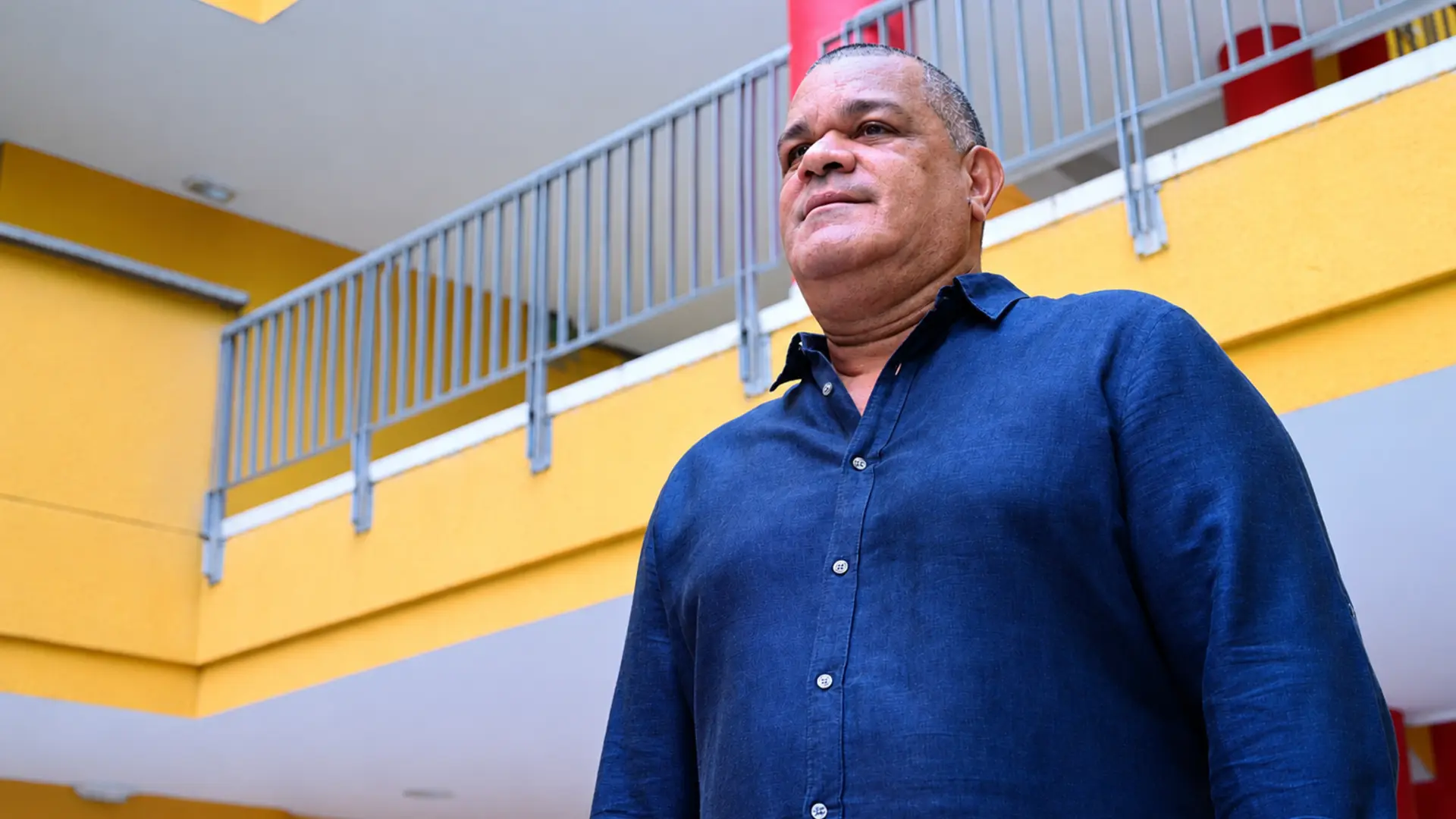 Rector de Uniguajira en el patio interior de un edificio institucional, visto desde un ángulo bajo. Viste una camisa azul oscura de manga larga y mira hacia el frente con expresión serena. Al fondo se observan balcones con barandas metálicas, paredes amarillas y columnas rojas que aportan color al entorno arquitectónico.