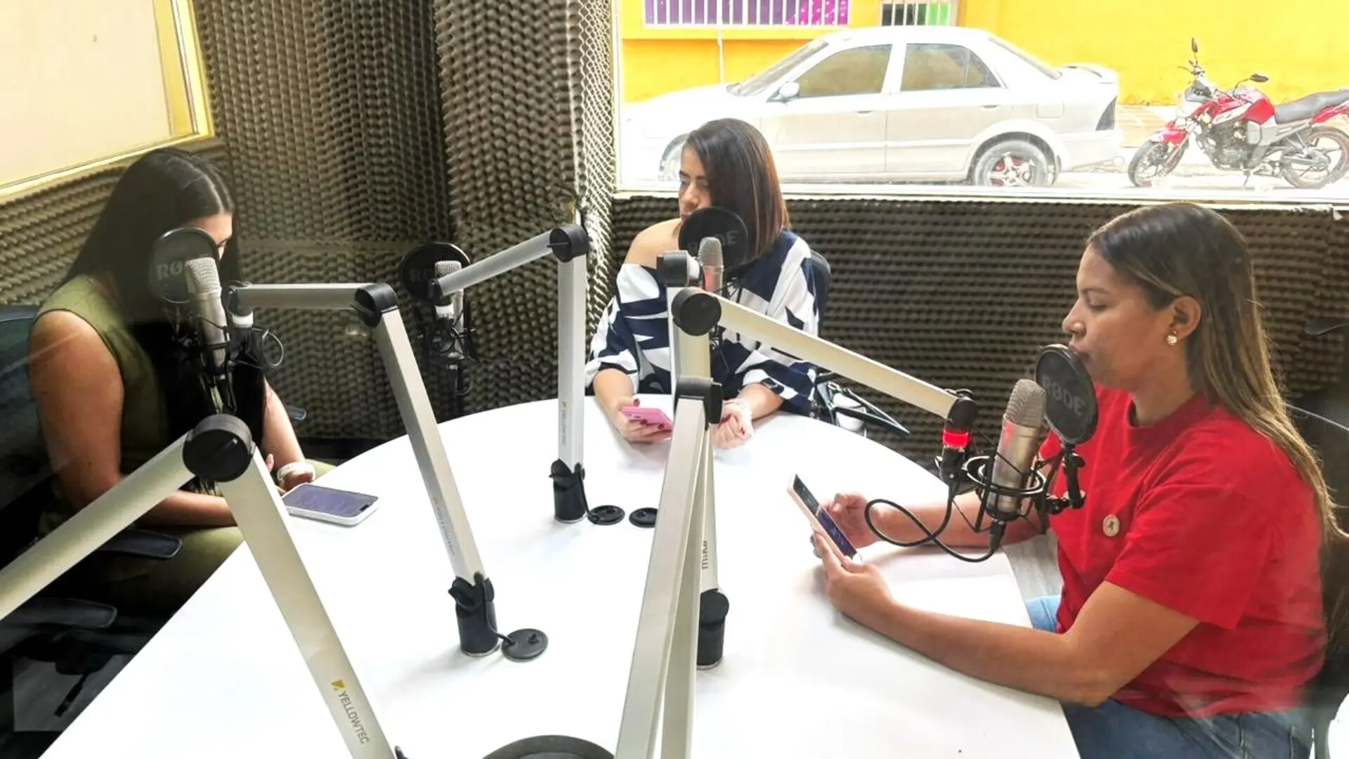 Tres mujeres participan en una conversación dentro de una cabina de radio, sentadas alrededor de una mesa con micrófonos profesionales. Una de ellas interviene mientras las otras escuchan y consultan sus dispositivos móviles. El estudio está recubierto con paneles acústicos y cuenta con una amplia ventana que permite observar el exterior