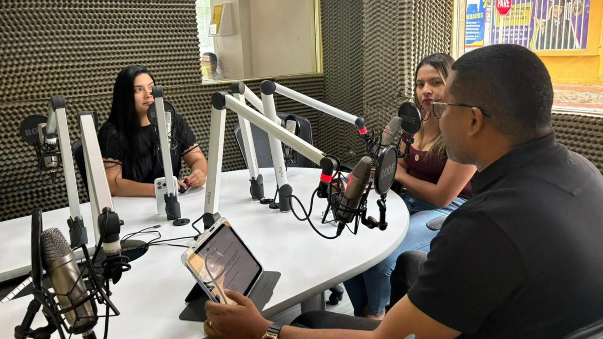Dos mujeres y un hombre participan en una conversación dentro de una cabina de radio, sentados alrededor de una mesa con micrófonos profesionales. El hombre dirige la conversación mientras sostiene una tableta electrónica, y las mujeres escuchan atentamente. El estudio está recubierto con paneles acústicos y tiene una ventana que permite la entrada de luz natural y la vista al exterior.