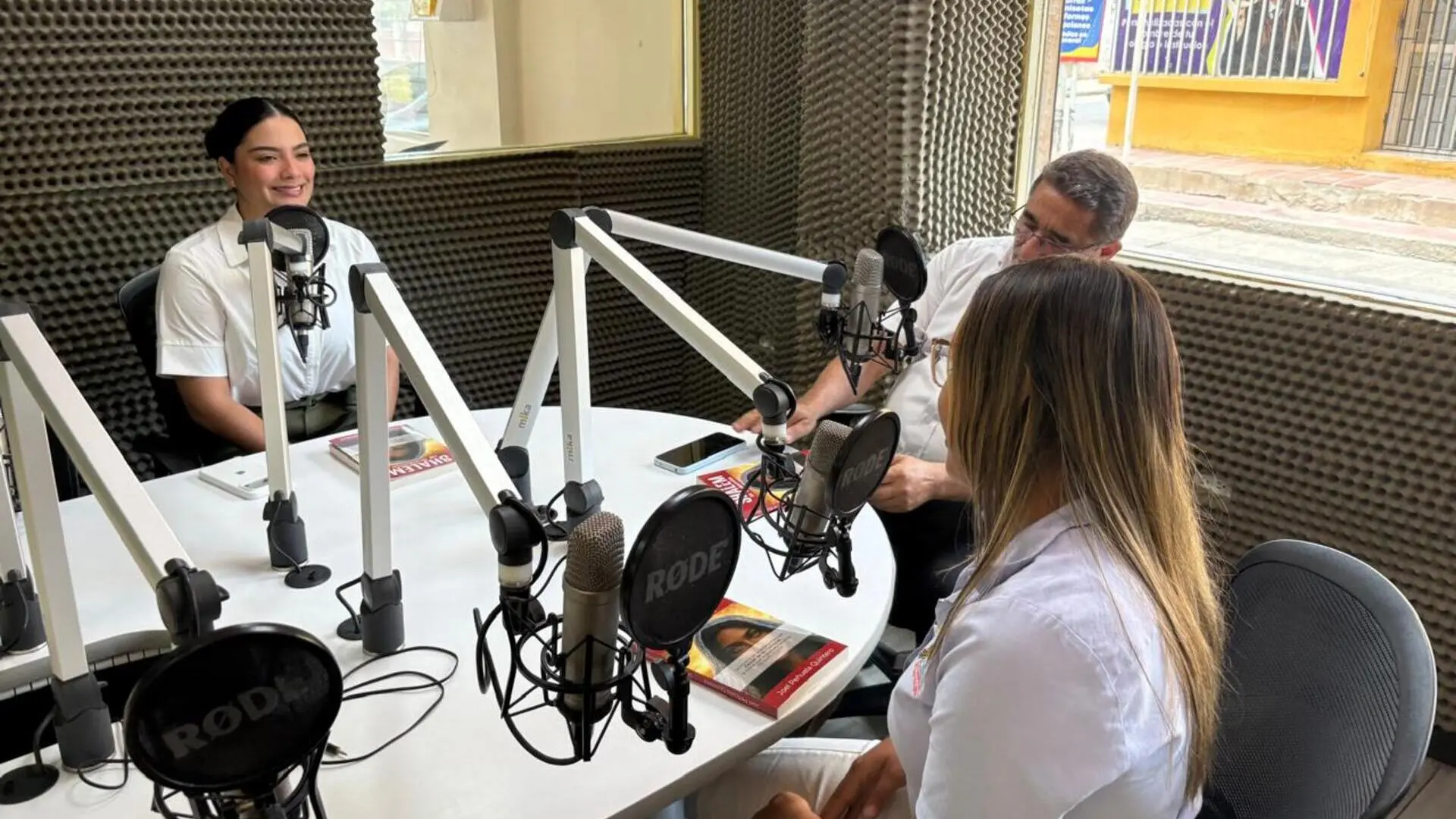 Dos mujeres y un hombre participan en una conversación dentro de una cabina de radio, sentados alrededor de una mesa con micrófonos profesionales. Una de las mujeres habla mientras los demás escuchan y consultan materiales de apoyo ubicados sobre la mesa. El estudio está recubierto con paneles acústicos y cuenta con una ventana que permite observar el exterior.