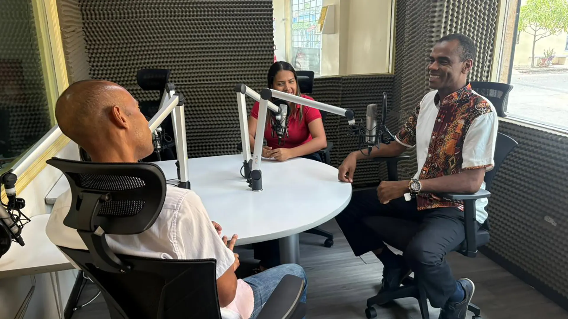 Tres personas participan en una conversación dentro de una cabina de radio profesional, sentadas alrededor de una mesa circular equipada con micrófonos de estudio. Una mujer y dos hombres intercambian ideas en un ambiente ameno y colaborativo. Los participantes muestran una actitud atenta y sonriente, reflejando una conversación dinámica y cercana.